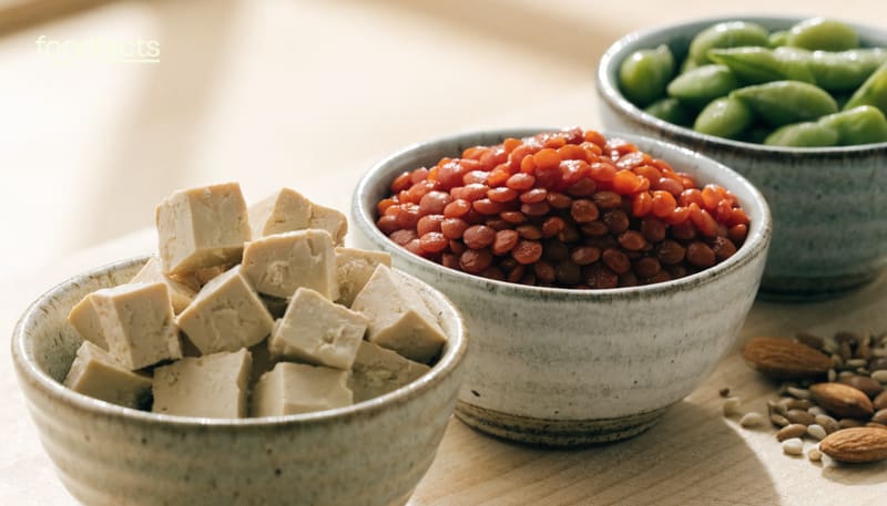 Three bowls of plant protein on a light wooden surface: cubed firm tofu, cooked red lentils and shelled edamame, with almonds and mixed seeds