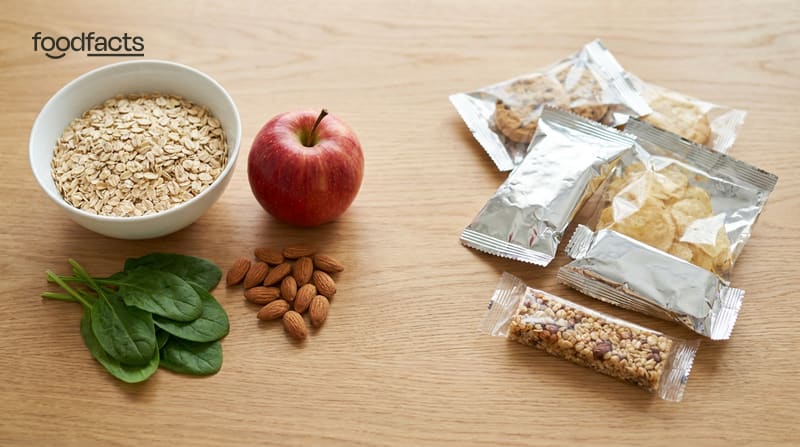 Whole foods — a bowl of oats, an apple, spinach and almonds — beside packaged ultra-processed snacks on a wooden table