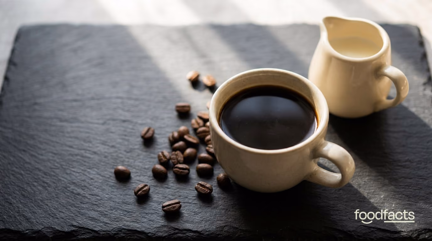 A cup of black coffee with a jug of milk and scattered coffee beans on a dark slate surface