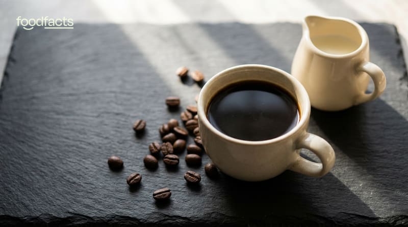 A cup of black coffee with a jug of milk and scattered coffee beans on a dark slate surface