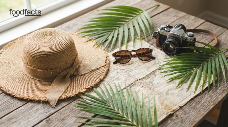 A straw sun hat, sunglasses, a film camera, a folded map and palm leaves on a wooden table