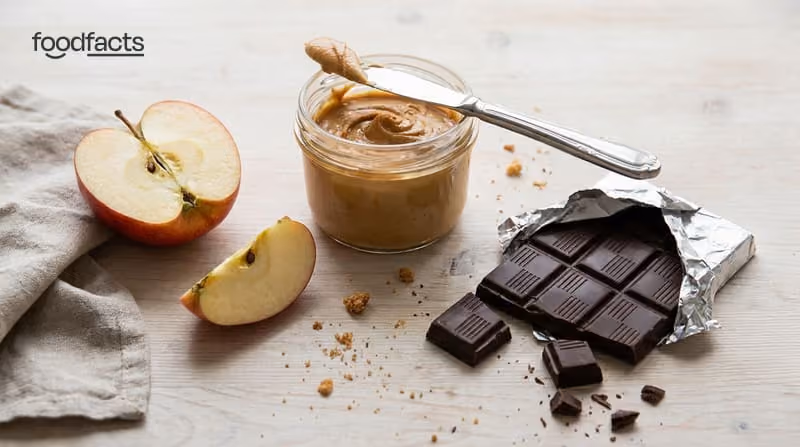 A sliced apple, a jar of peanut butter and a chocolate bar side by side on a table