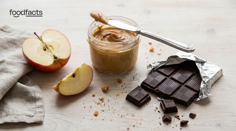 A sliced apple, a jar of peanut butter and a chocolate bar side by side on a table