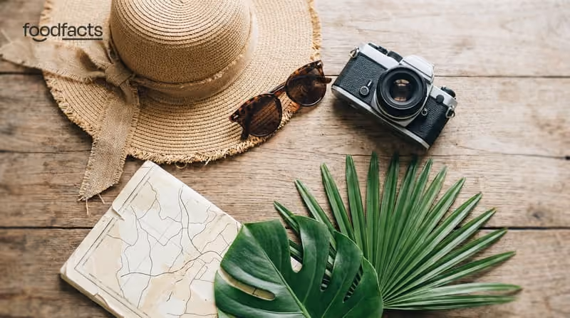 A straw sun hat, sunglasses, a film camera, a folded map and palm leaves on a wooden table