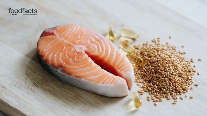 Salmon fillet and sardines on ice beside bowls of flaxseeds, chia seeds and walnuts, with omega-3 capsules and seaweed.