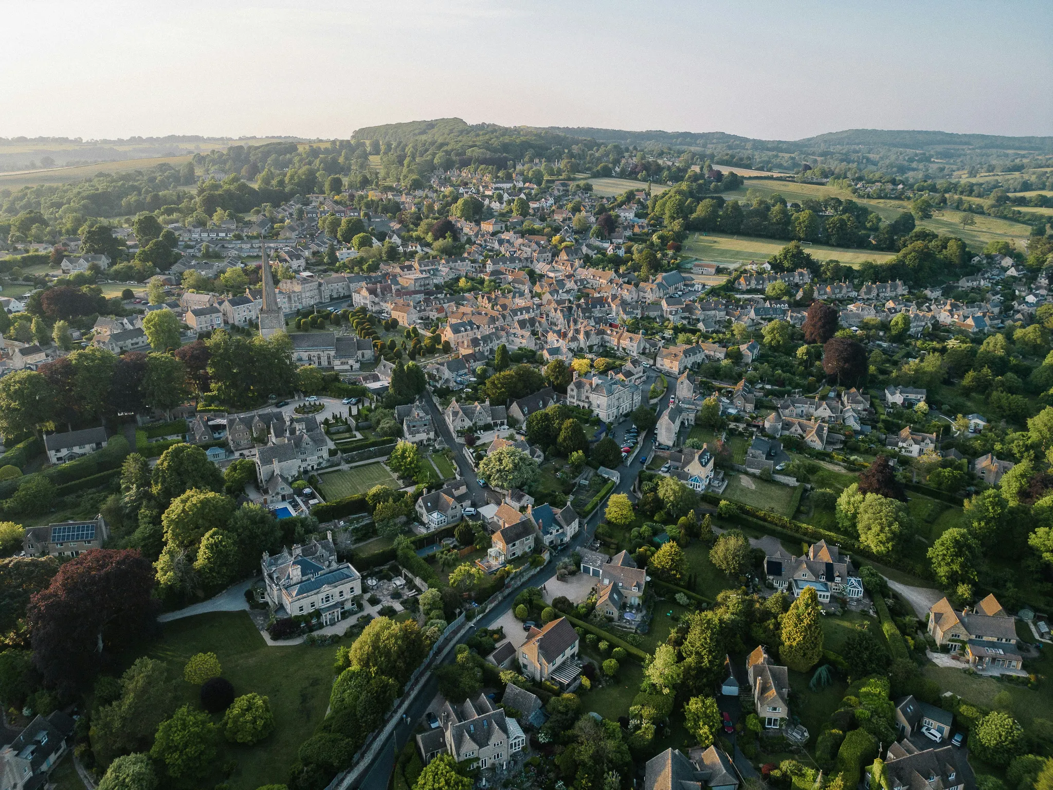 Aerial view of a quaint village with stone houses, a church with a tall spire, and green trees surrounding residential areas.