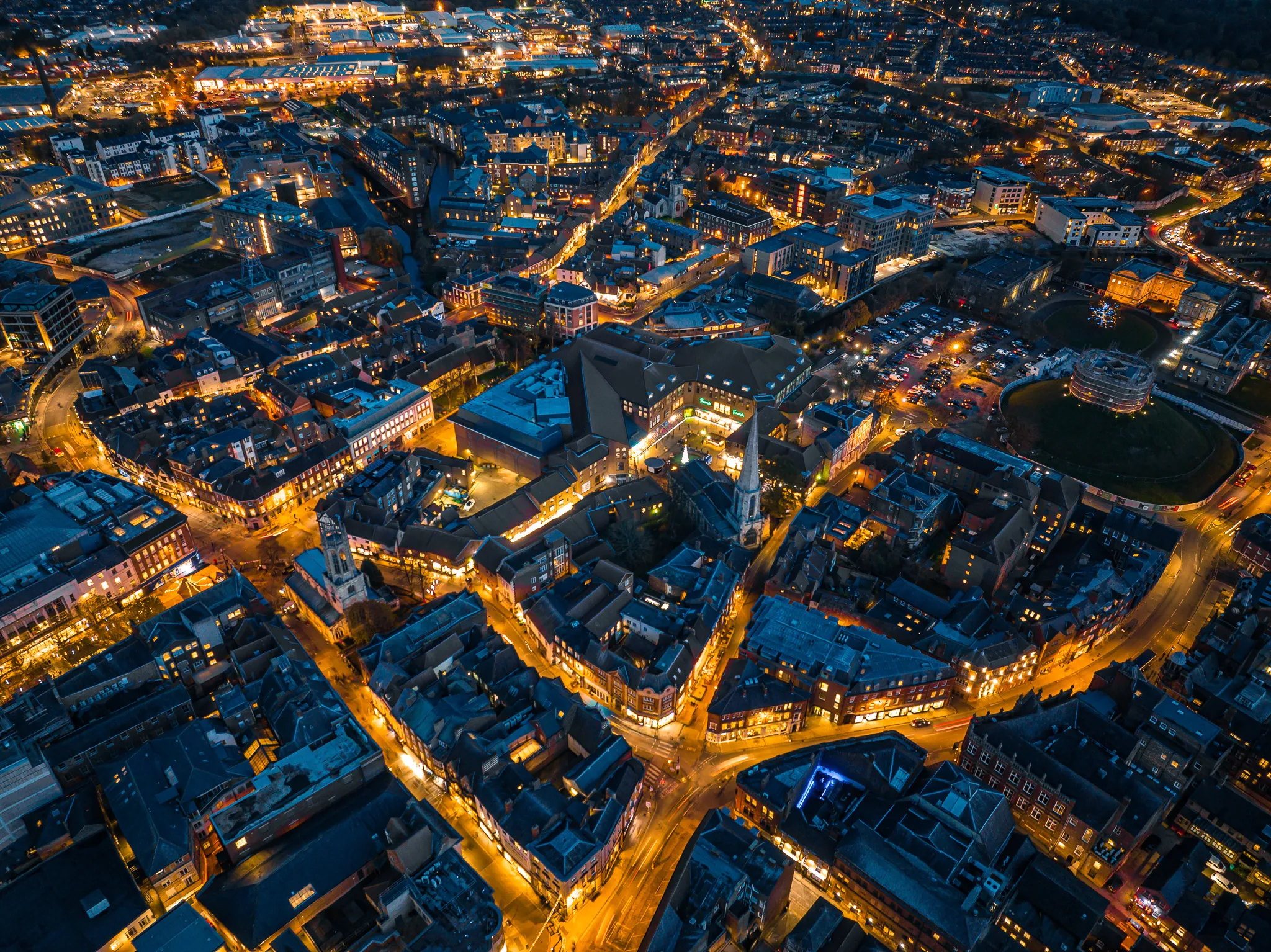 Aerial view of a city at night with illuminated streets, buildings, parking lots, and a park area with a circular mound.