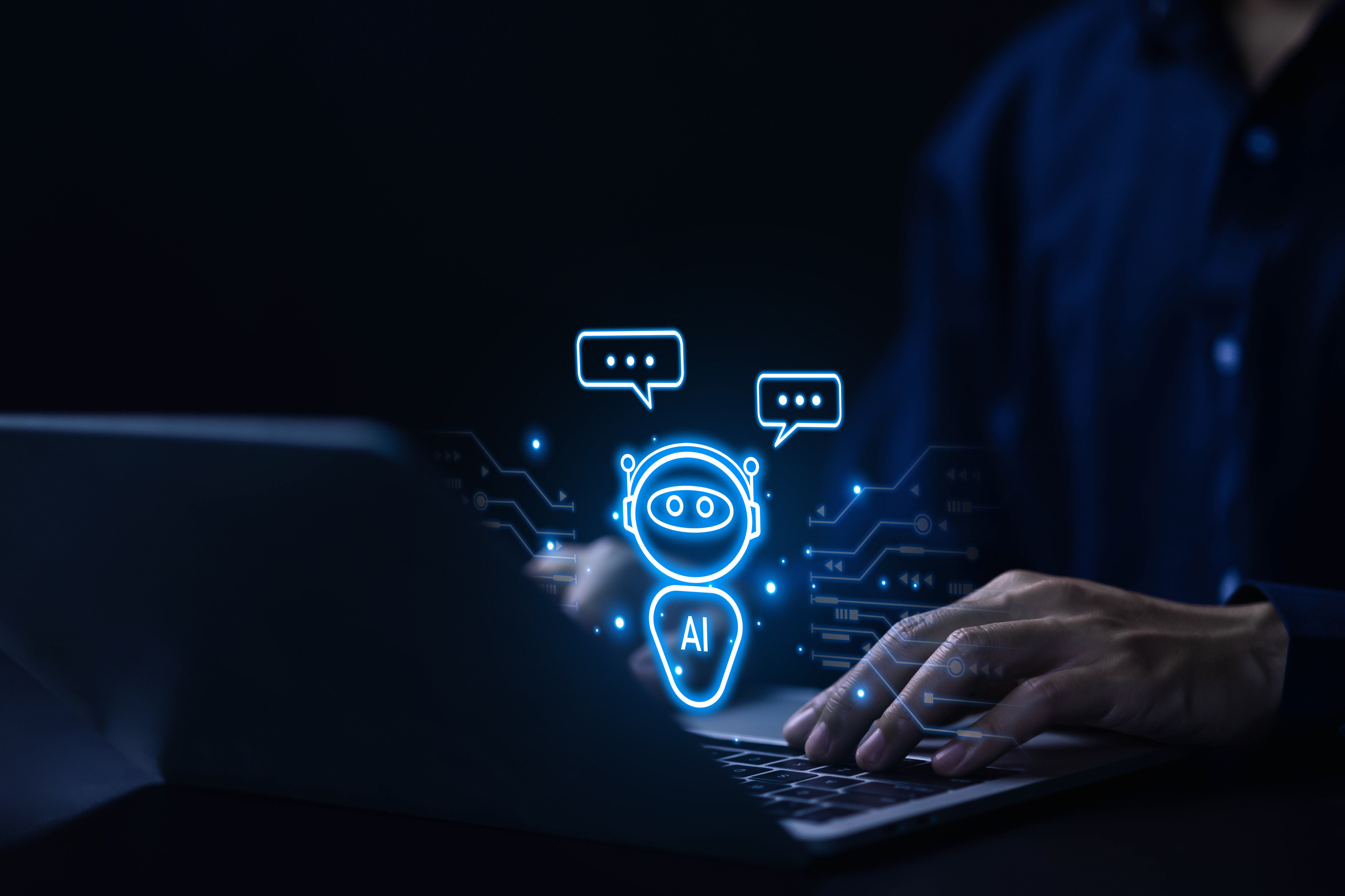 Dark, low-light image of a person sitting at a desk and typing on a laptop. A glowing, blue digital overlay of an AI chatbot icon—a robot head with 'AI' written below it and chat bubbles above it—is projected onto the center of the image, symbolizing artificial intelligence or automation.