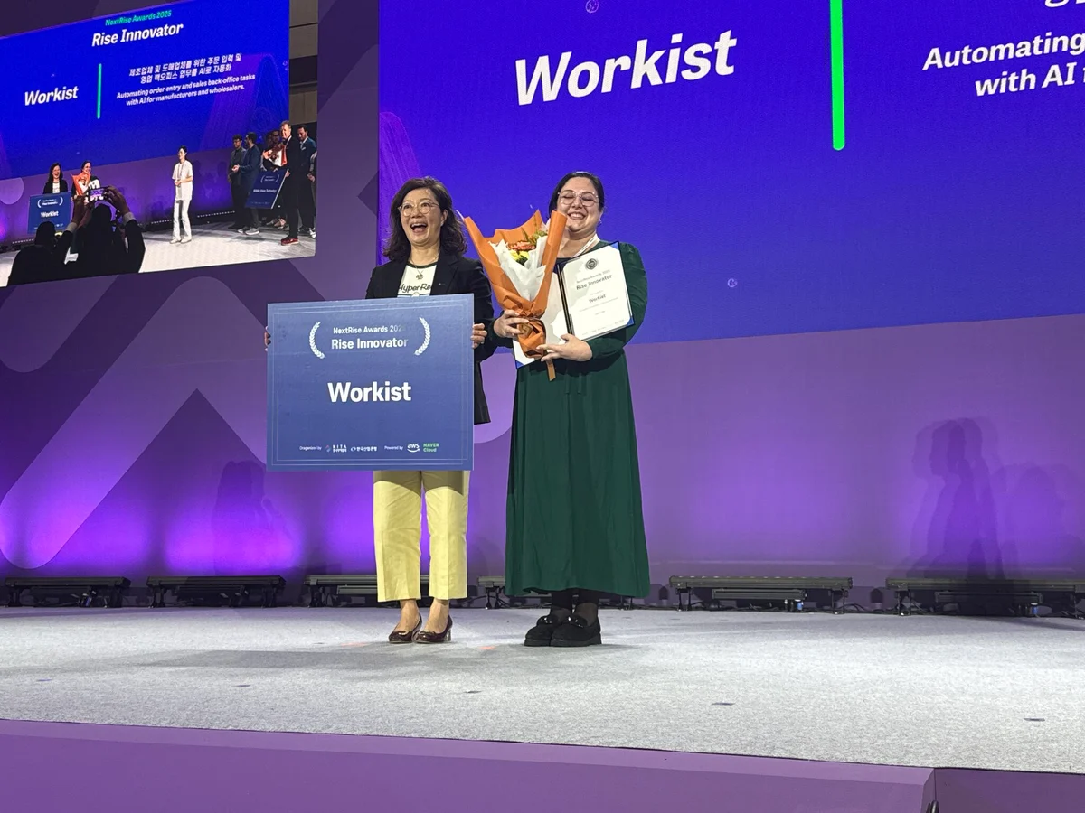 Two women standing on a stage, one on the left holding a large 'Workist Rise Innovator Award 2023' sign, and the woman on the right holding flowers and a certificate, in front of a screen displaying 'Workist'