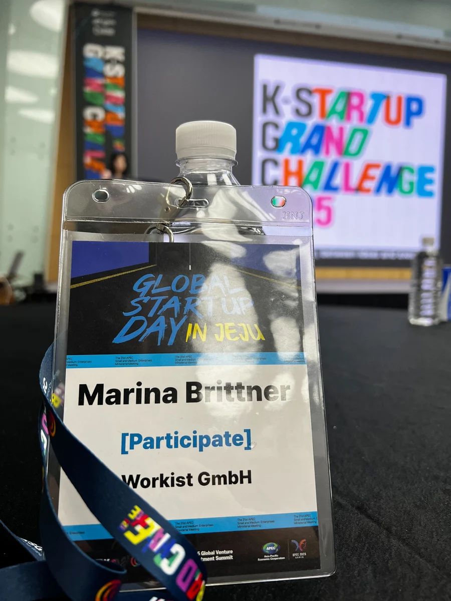 Close-up of a participant ID badge and blue lanyard lying on a dark surface. A blurred screen in the background displays the text 'K-STARTUP GRAND CHALLENGE'.