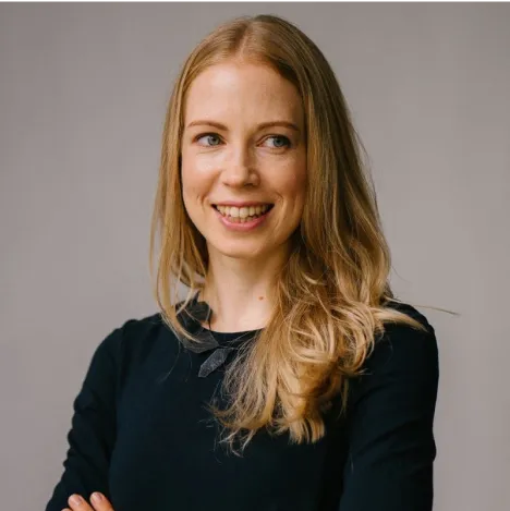 Femme souriante aux cheveux longs blonds, vêtue d'un haut noir, posant devant un fond neutre.