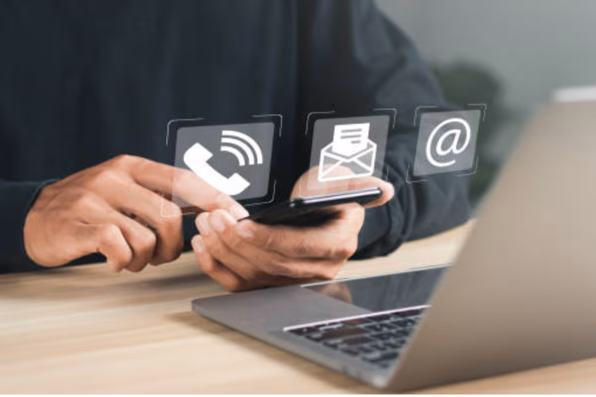 Personne utilisant un smartphone devant un ordinateur portable avec des icônes flottantes de téléphone, enveloppe et symbole arobase.