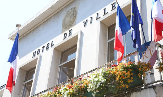 Façade d'un bâtiment officiel avec l'inscription 'HOTEL DE VILLE', des drapeaux français et européens, et des jardinières fleuries.