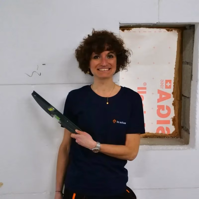 Une femme souriante tenant un niveau à bulle devant un mur blanc avec une ouverture rectangulaire.
