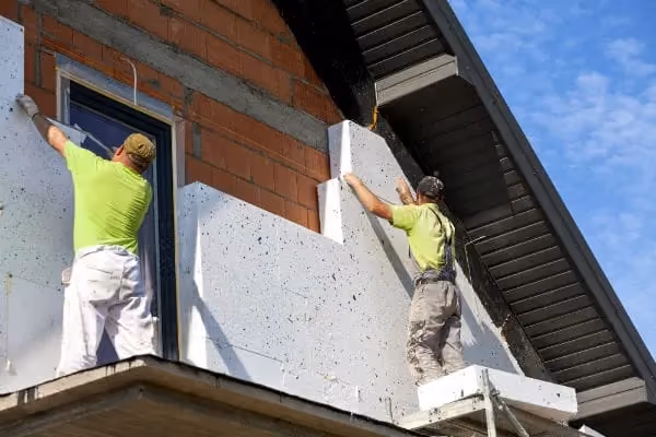 Deux ouvriers installent des panneaux d'isolation blanche sur la façade en briques d'une maison sous un toit en pente.
