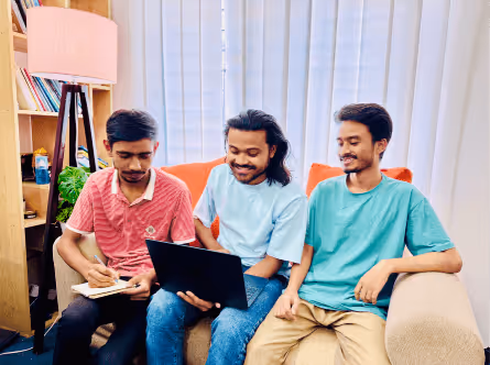 Three young men sitting on a beige couch, one writing in a notebook, while another holds a laptop, engaged in discussion.