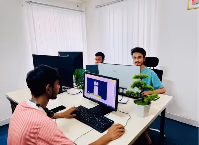 Three men working at computers around a white office desk with small potted plants.