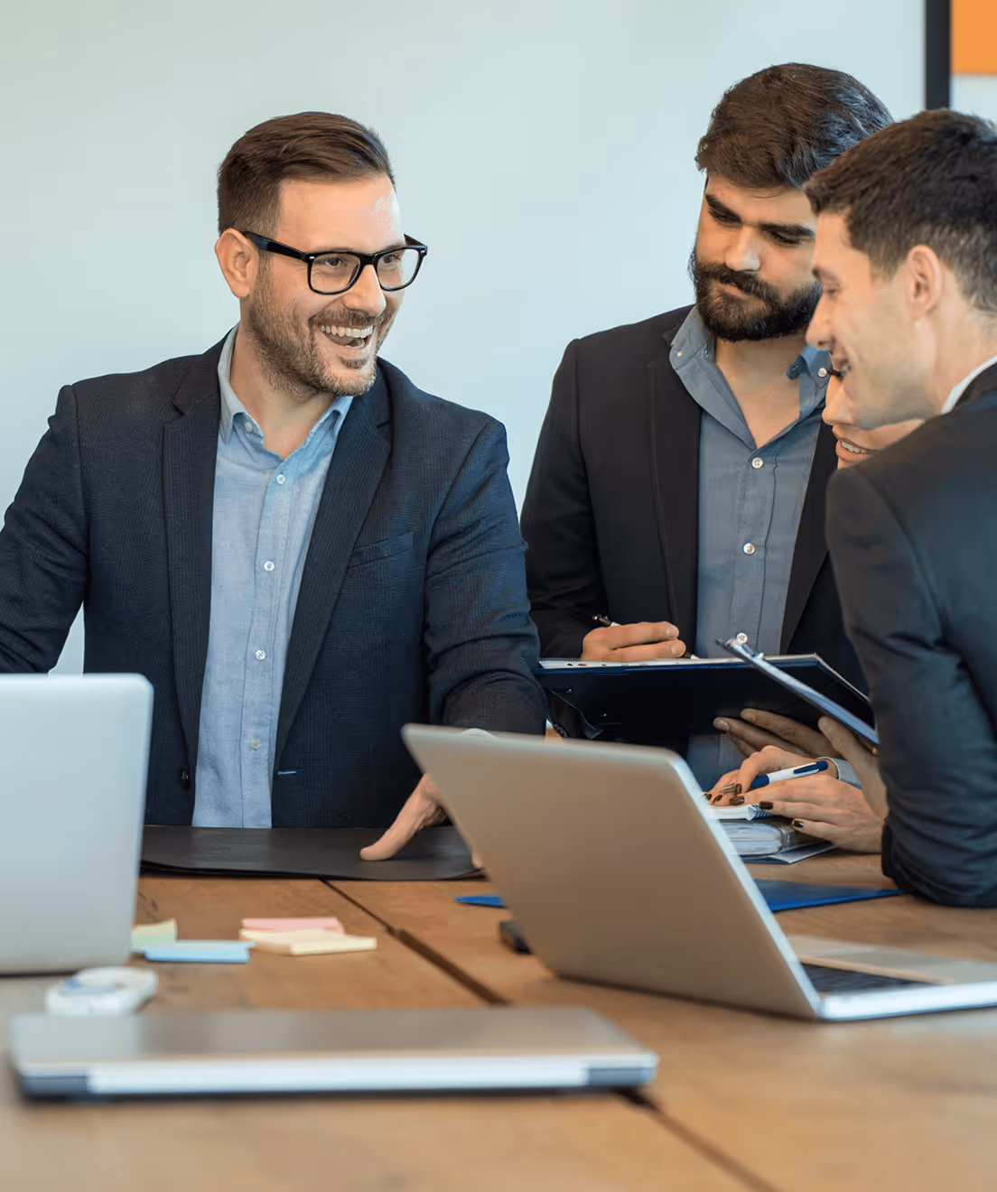 Four businesspeople in suits smiling and discussing work with laptops and documents on a table.