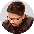 Young man with short dark hair and glasses wearing a black shirt looking downward thoughtfully.