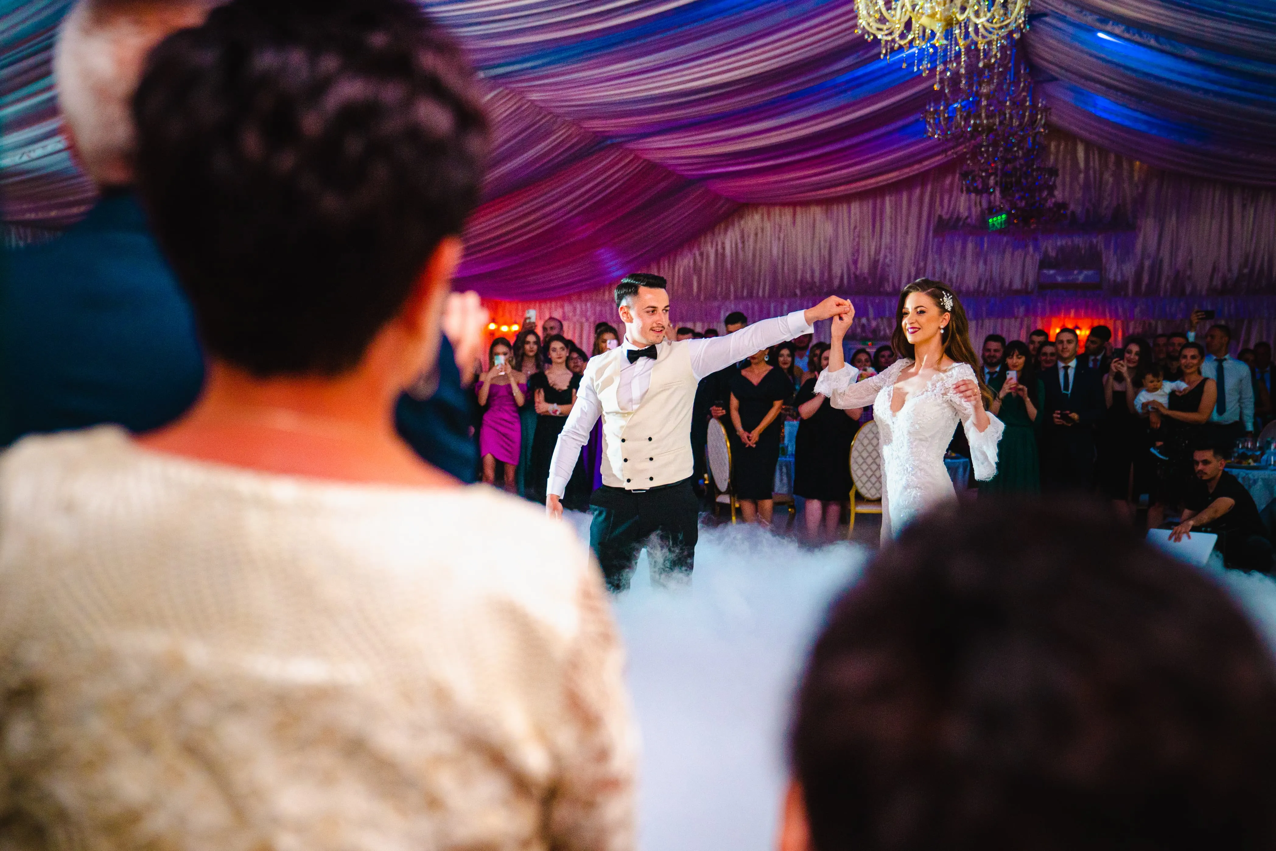 Fotografie de nuntă realizată de Flaviu Studio cu un cuplu de miri dansând primul dans în fața invitaților într-o sală de festivități elegant decorată. Mireasa poartă o rochie albă cu dantelă și mirele este îmbrăcat într-o vestă albă cu papion negru. Atmosfera este iluminată festiv, iar podeaua este acoperită cu o ceață ușoară, creând un moment magic.
