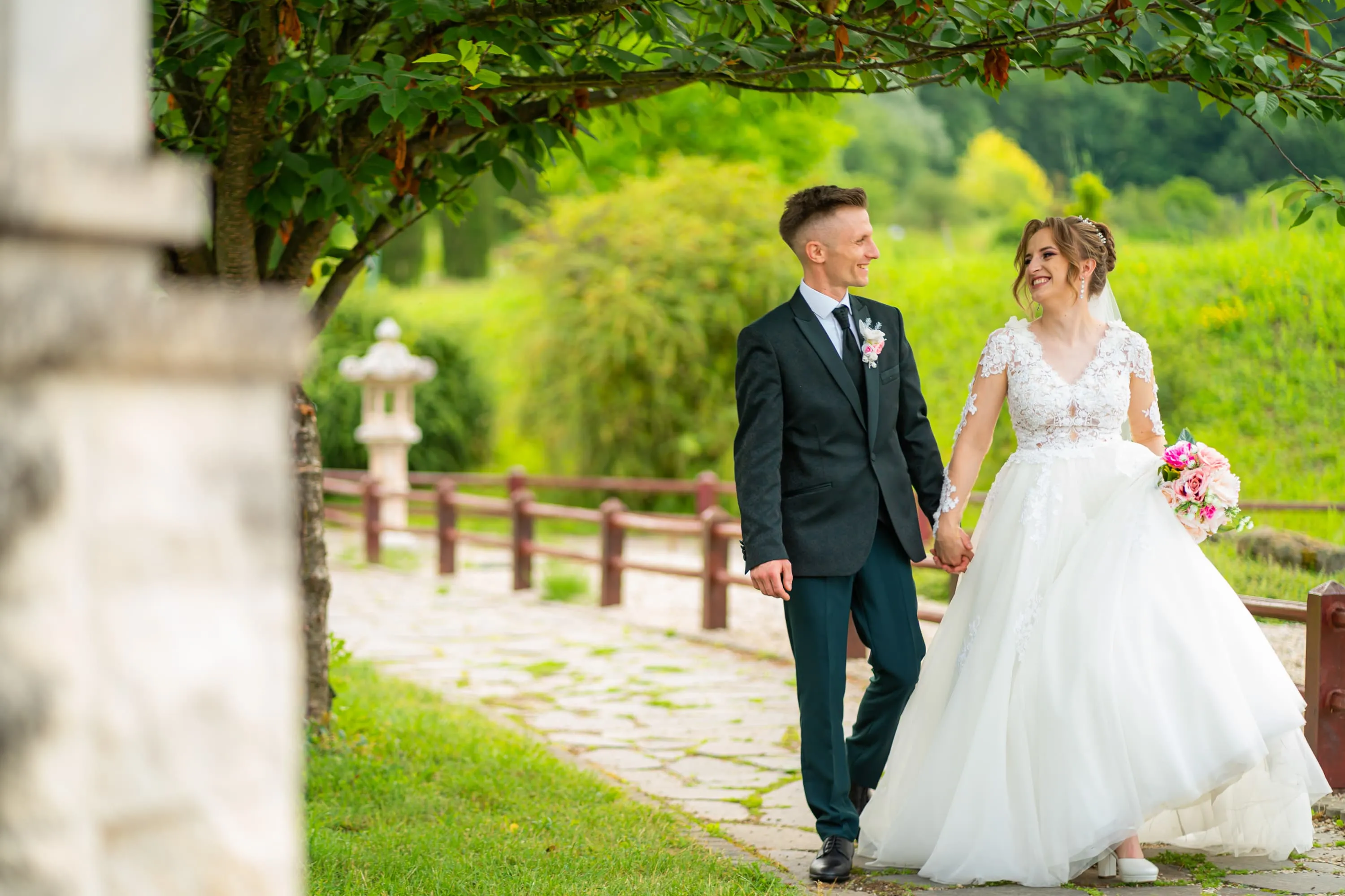 Cuplu de miri plimbându-se pe o alee înconjurată de verdeață, la nunta lor. Mireasa poartă o rochie albă cu dantelă și ține un buchet de flori roz, iar mirele este îmbrăcat într-un costum negru. Fotografie de nuntă realizată de Flaviu Studio.