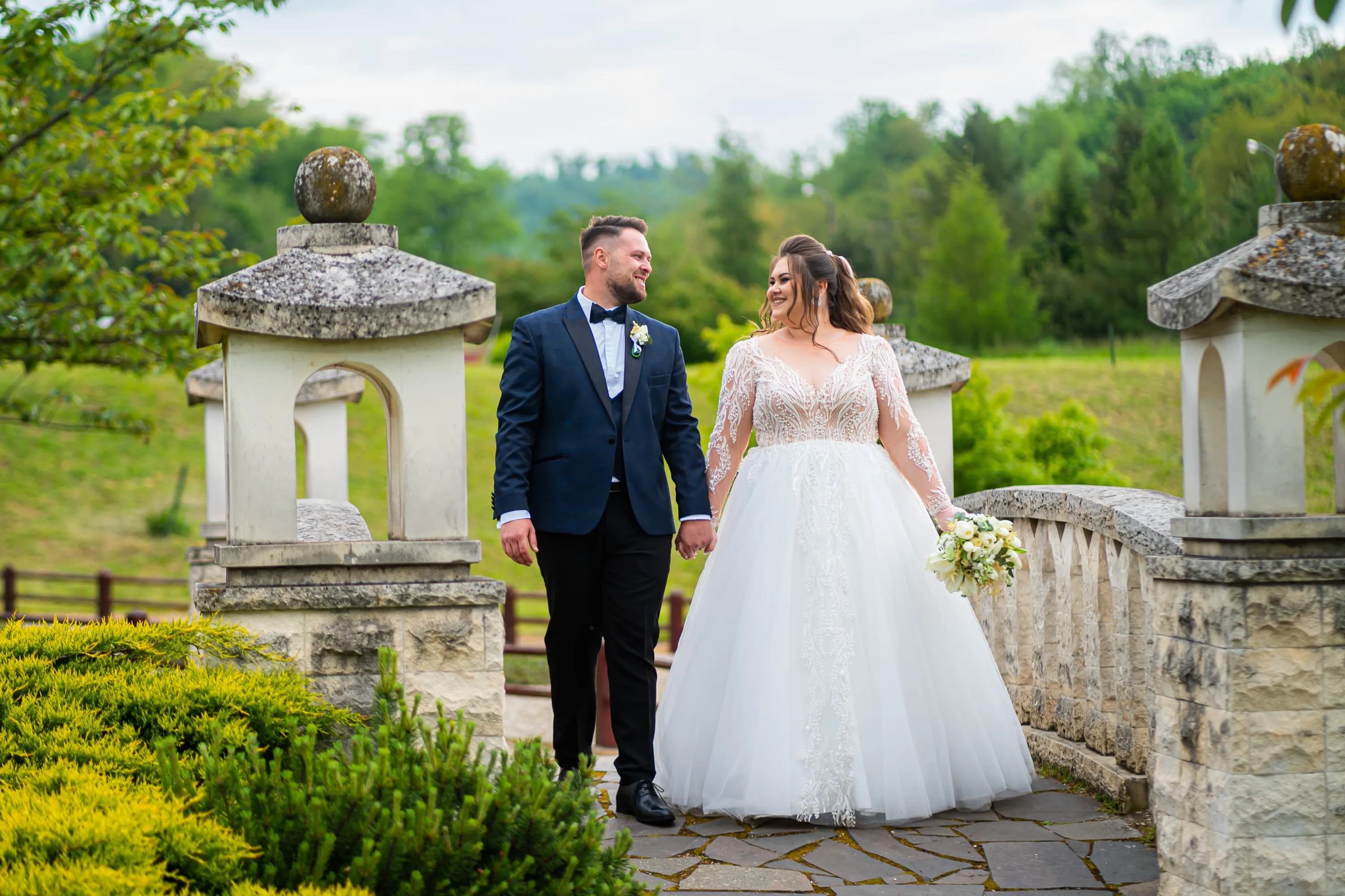 Cuplu de miri plimbându-se pe un pod de piatră, înconjurat de verdeață, la nunta lor. Mireasa poartă o rochie albă cu dantelă și ține un buchet de flori, iar mirele este îmbrăcat într-un costum negru. Fotografie de nuntă realizată de Flaviu Studio.