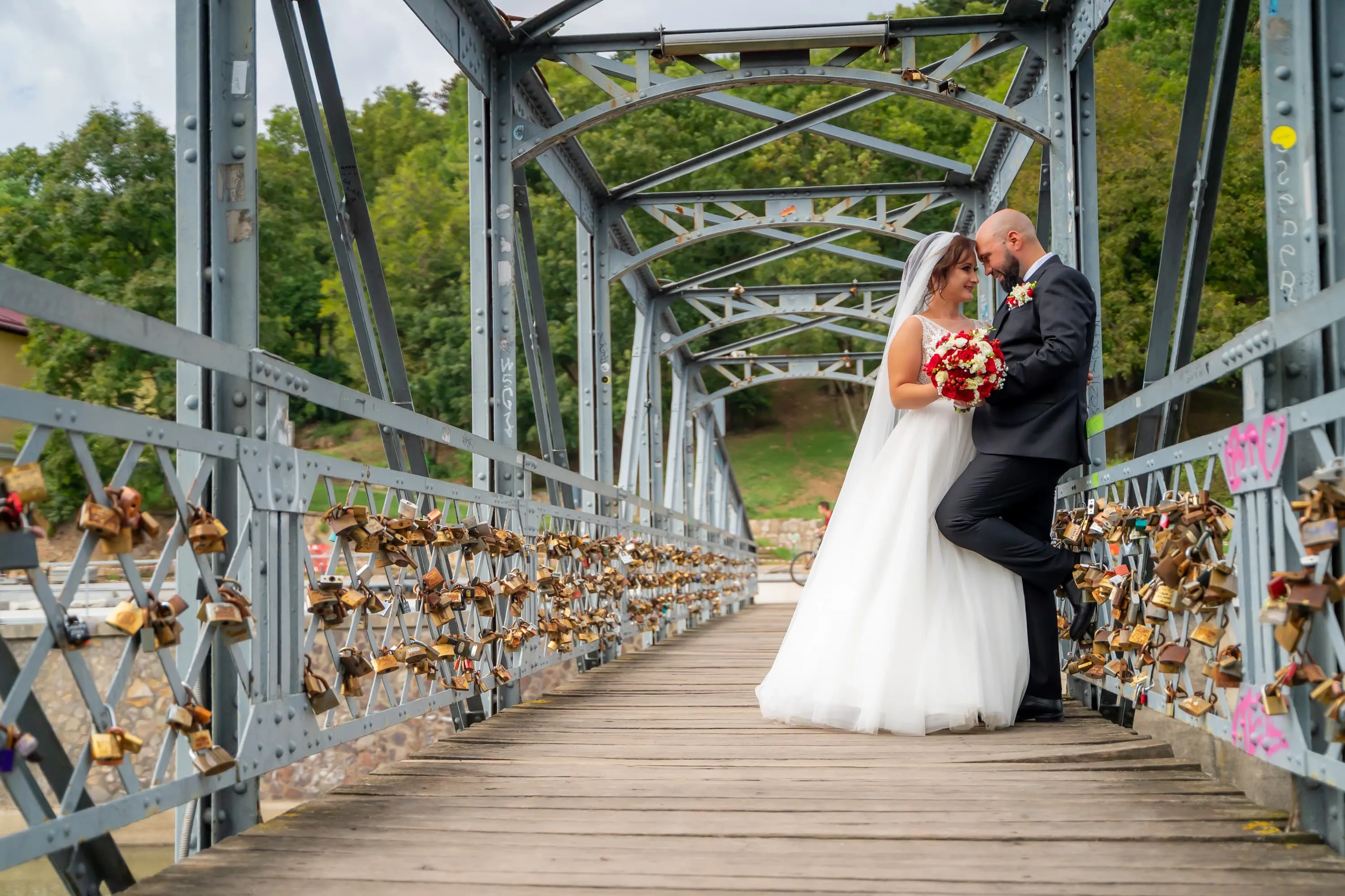 Cuplu de miri pe un pod decorat cu lacăte de dragoste, la nunta lor. Mireasa poartă o rochie albă și ține un buchet roșu, iar mirele este îmbrăcat într-un costum negru. Fotografie de nuntă realizată de Flaviu Studio.