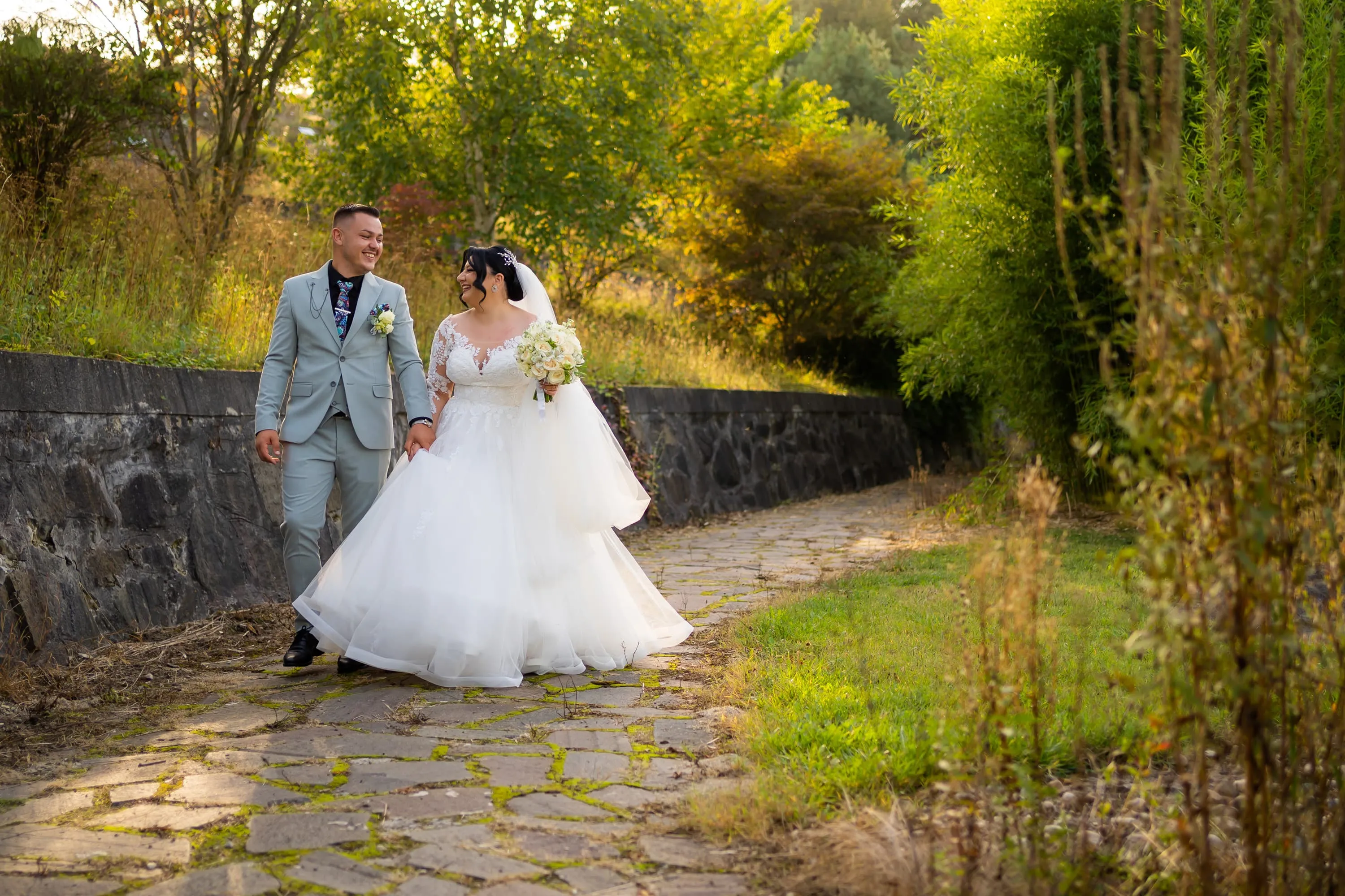 Cuplu de miri plimbându-se pe o alee pietruită, înconjurată de verdeață, la nunta lor. Mireasa poartă o rochie albă cu voal și ține un buchet de flori, iar mirele este îmbrăcat într-un costum gri deschis. Fotografie de nuntă realizată de Flaviu Studio.