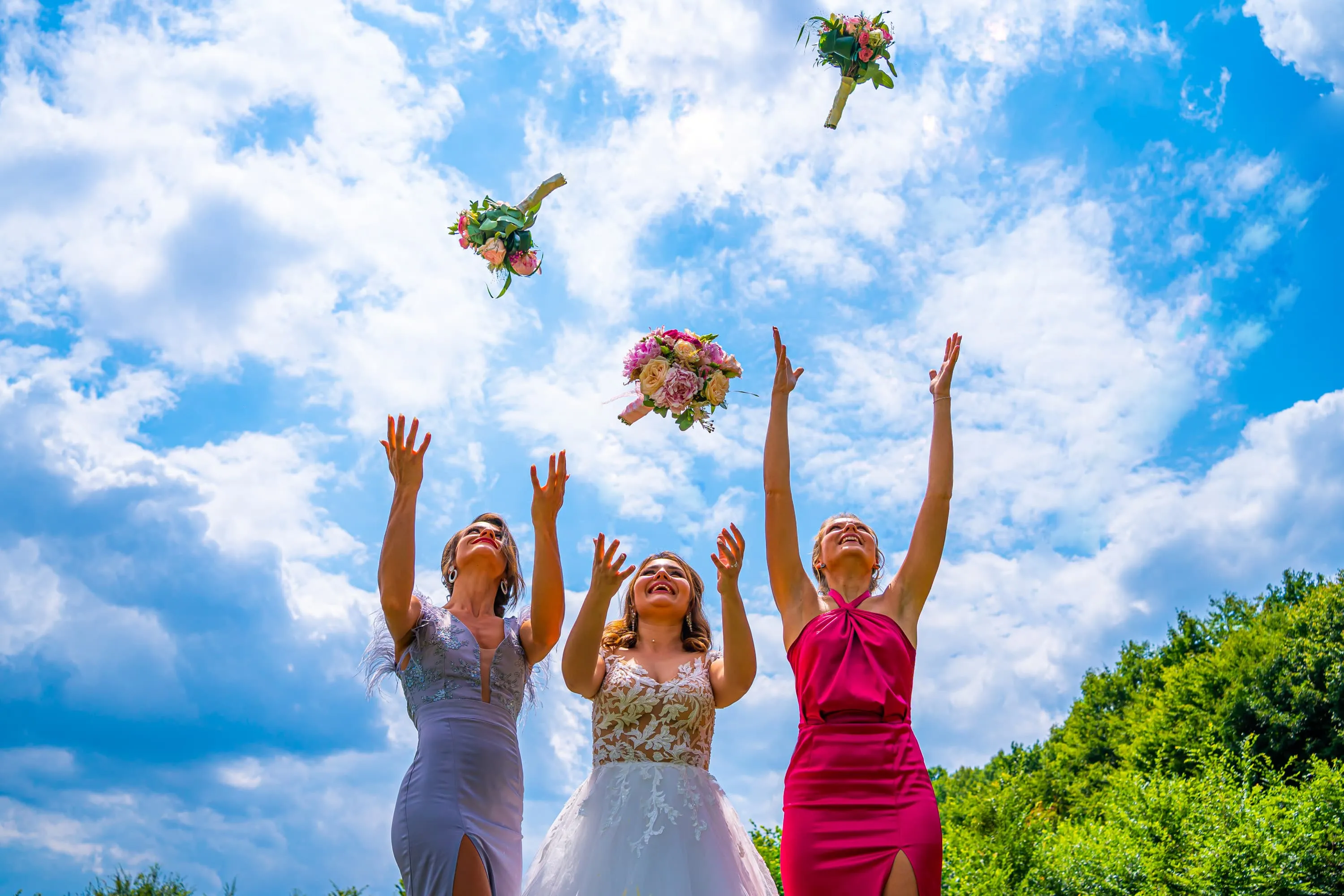 Fotografie de nuntă realizată de Flaviu Studio cu mireasa și două domnișoare de onoare aruncând buchetele în aer pe un cer senin și albastru. Mireasa poartă o rochie albă cu detalii de dantelă, în timp ce domnișoarele de onoare sunt îmbrăcate în rochii elegante, una în roz și cealaltă în mov deschis. Momentul surprinde bucuria și veselia evenimentului într-un cadru natural.