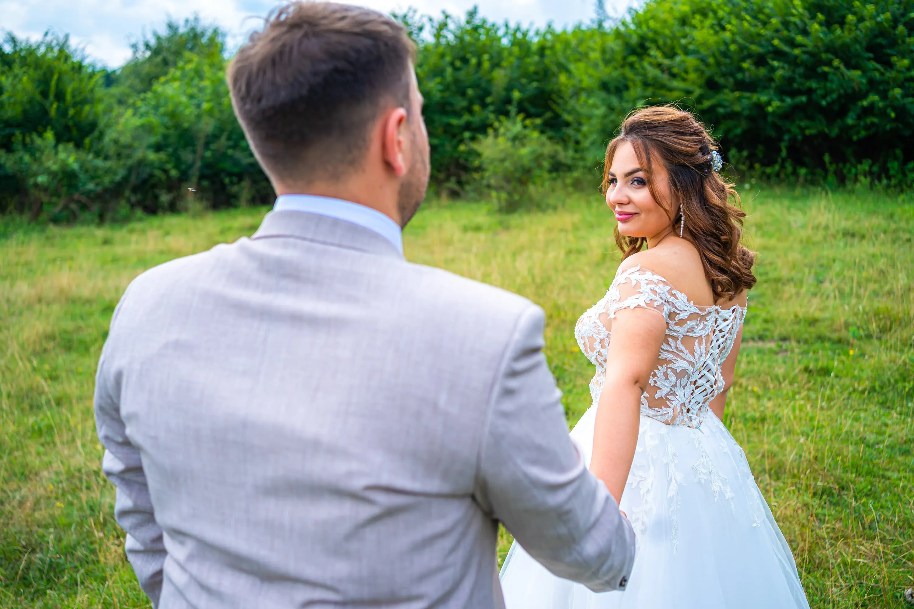 Fotografie de nuntă realizată de Flaviu Studio cu mireasa și mirele ținându-se de mână într-o pajiște verde. Mireasa, purtând o rochie albă cu detalii florale de dantelă, privește zâmbitoare peste umăr către cameră, în timp ce mirele, îmbrăcat într-un costum deschis la culoare, îi întinde mâna. Fundalul natural creează o atmosferă romantică și idilică.