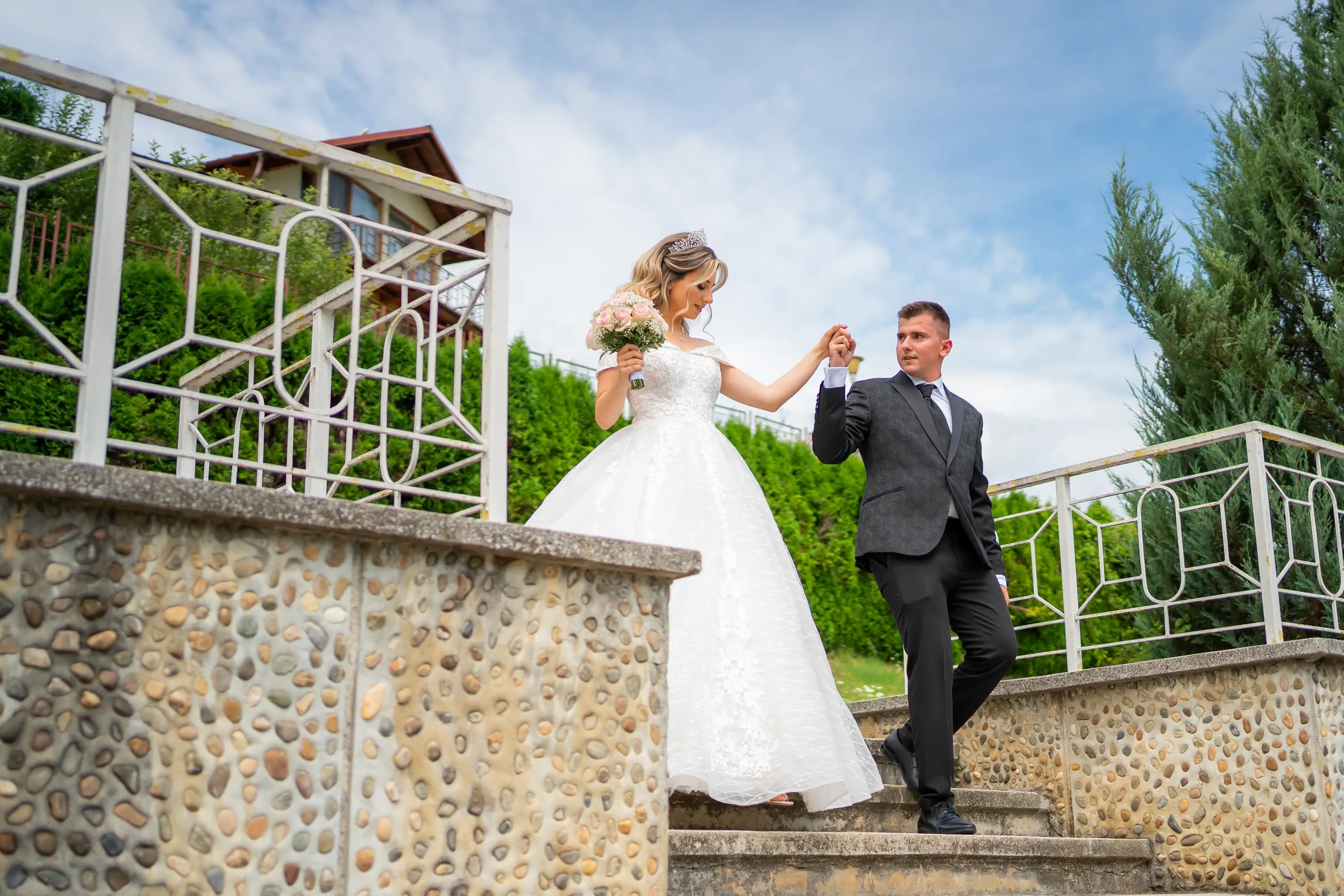 Cuplu de miri coborând treptele unei clădiri, la nunta lor. Mireasa poartă o rochie albă cu buchet de flori roz, iar mirele este îmbrăcat într-un costum negru. Fotografie de nuntă realizată de Flaviu Studio.