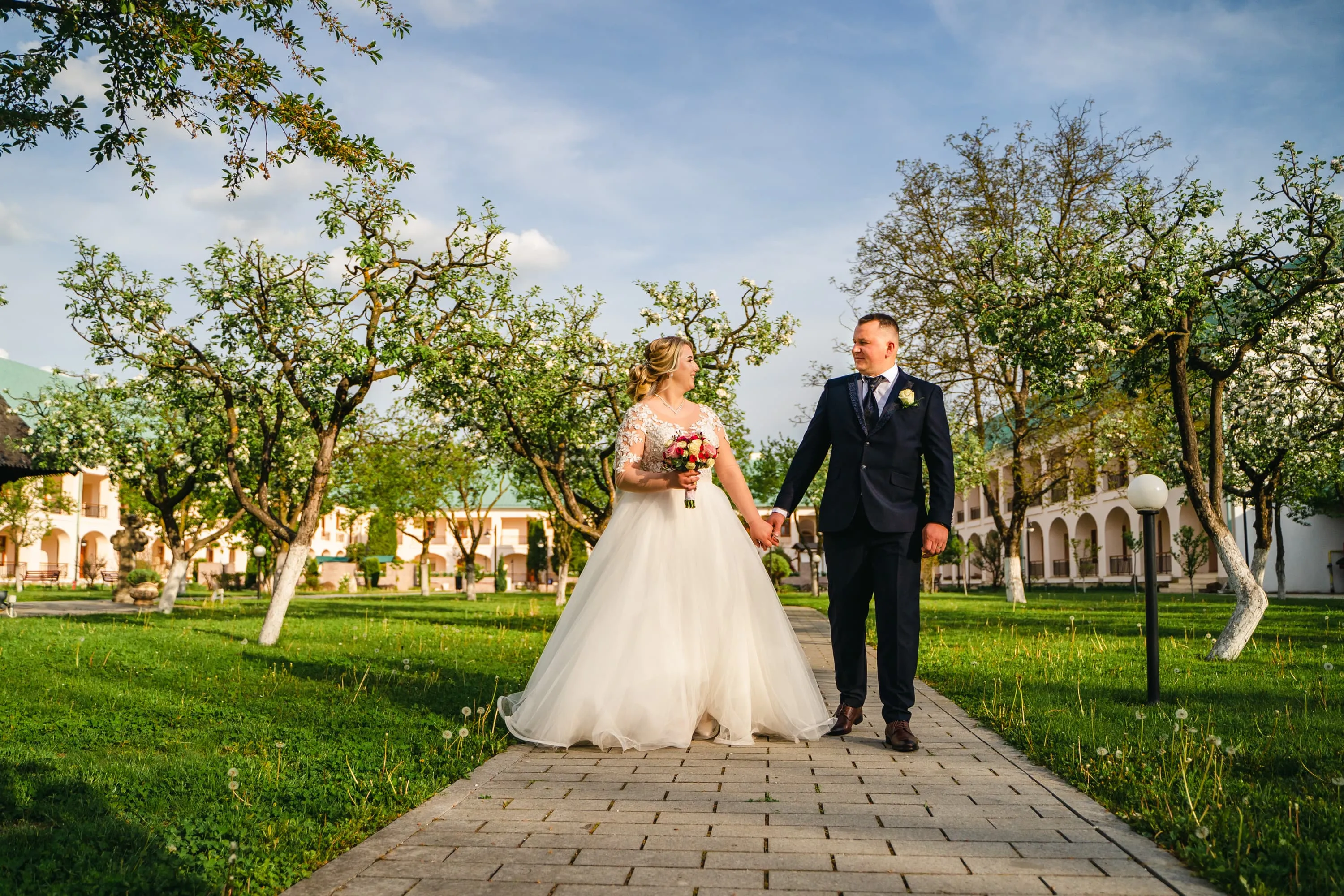 Cuplu de miri plimbându-se printr-o livadă înflorită, la nunta lor. Mireasa poartă o rochie albă și ține un buchet de flori, iar mirele este îmbrăcat într-un costum negru. Fotografie de nuntă realizată de Flaviu Studio.