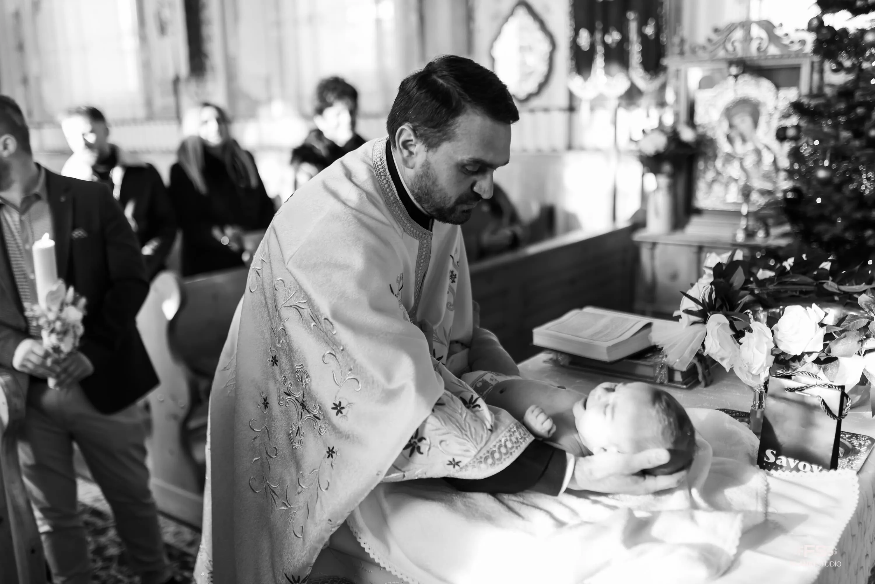 Preot ținând un bebeluș pe masa de botez în timpul ceremoniei religioase, cu familia și invitații privindu-i în fundal. Fotografie alb-negru realizată de Flaviu Studio.