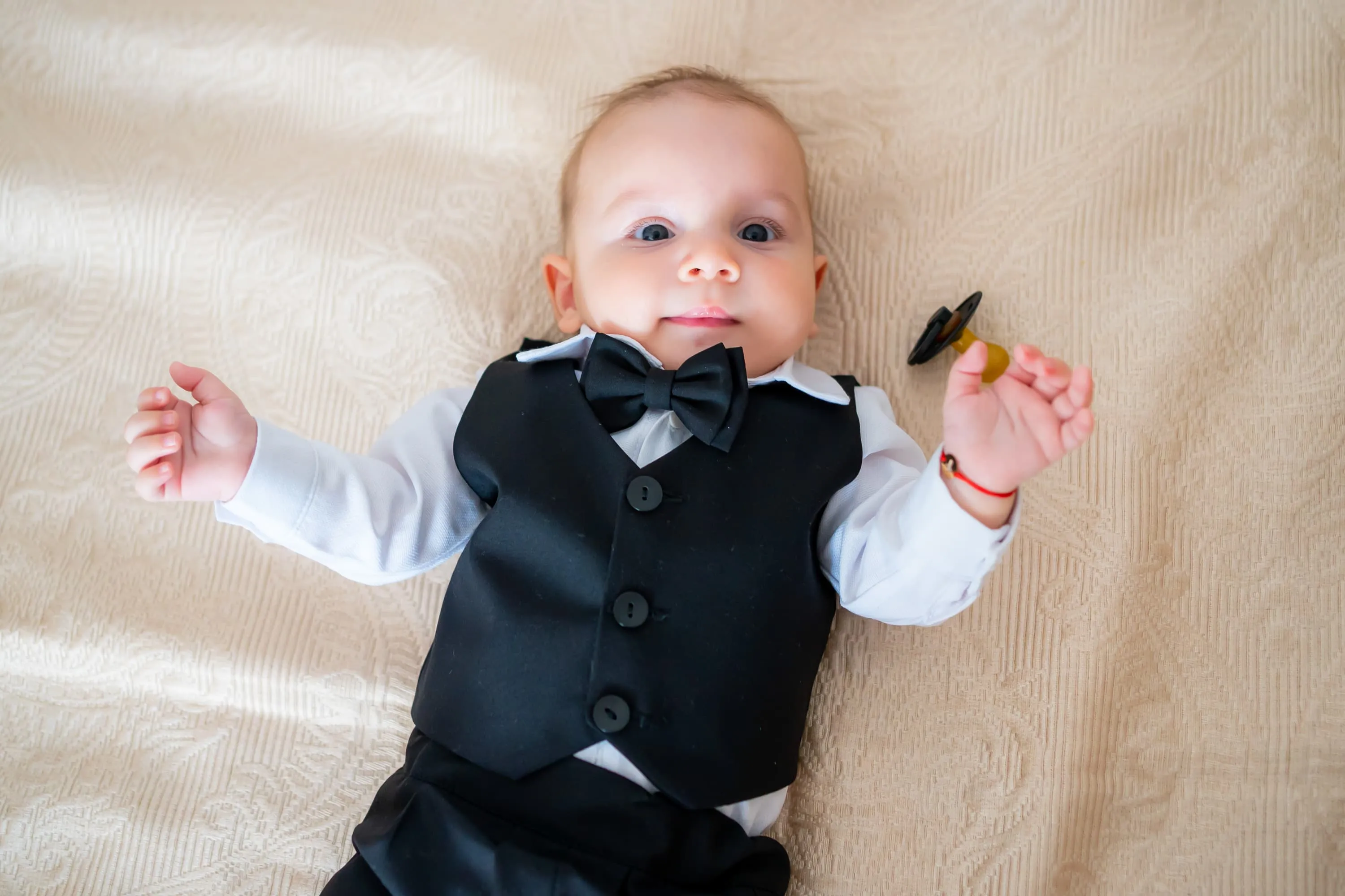 Bebeluș îmbrăcat într-un costum elegant negru, cu papion, stând pe spate pe un pat cu așternuturi deschise la culoare. Bebelușul are ochii mari și expresia feței este curioasă. Fotografie realizată de Flaviu Studio.