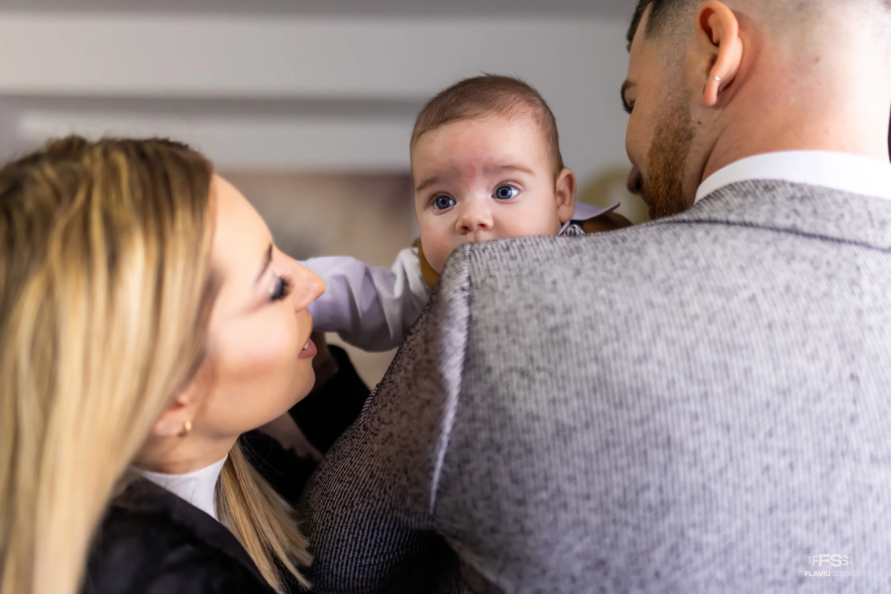 Bebeluș numit Eduard-Nicolas ținut în brațe de tatăl său, în timp ce mama privește zâmbitoare. Bebelușul este îmbrăcat elegant pentru botez. Fotografie realizată de Flaviu Studio.