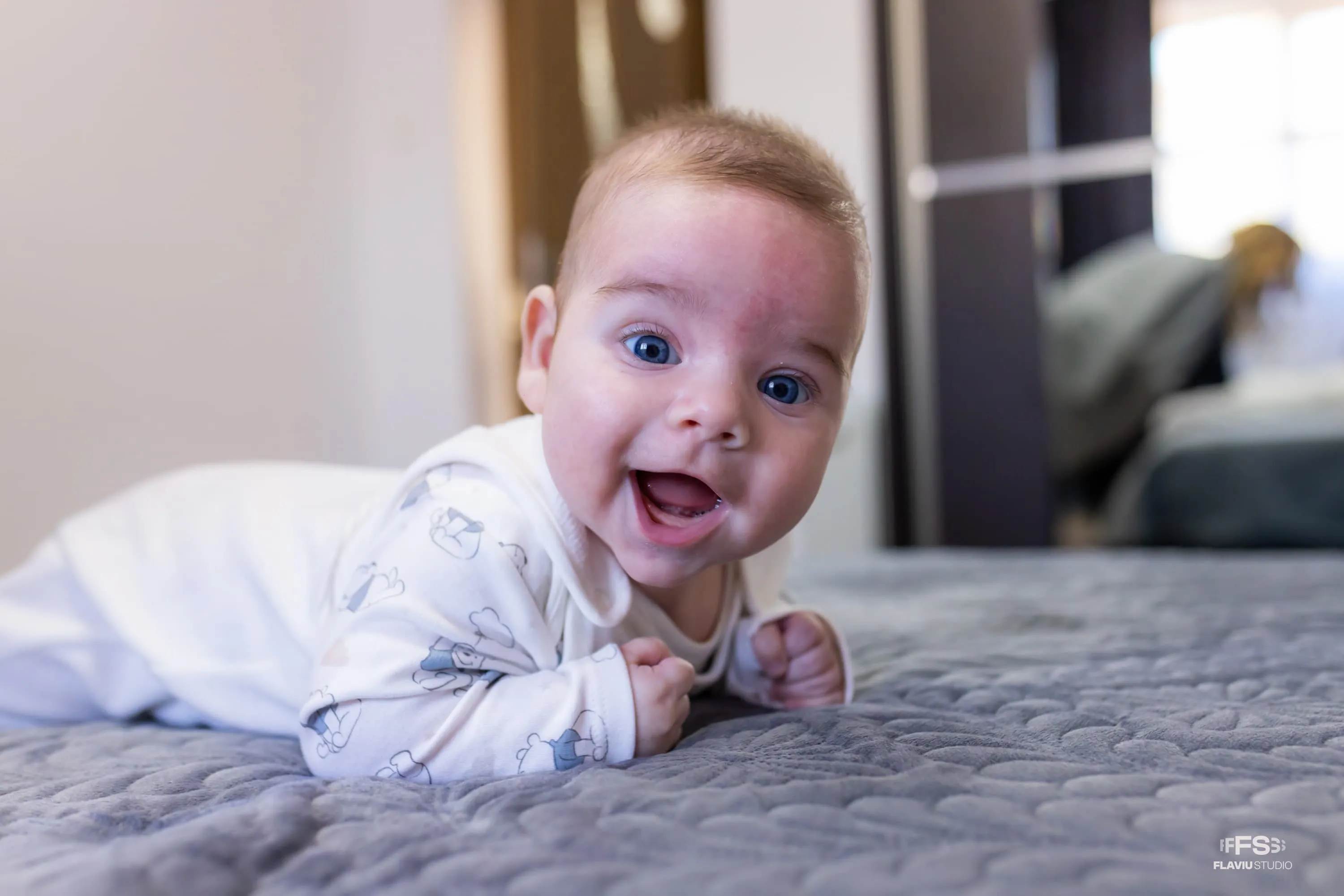 Bebeluș zâmbitor numit Eduard-Nicolas, îmbrăcat într-un costum alb cu desene de ursuleți, stând pe burtă pe un pat gri. Fotografie realizată de Flaviu Studio la un botez.