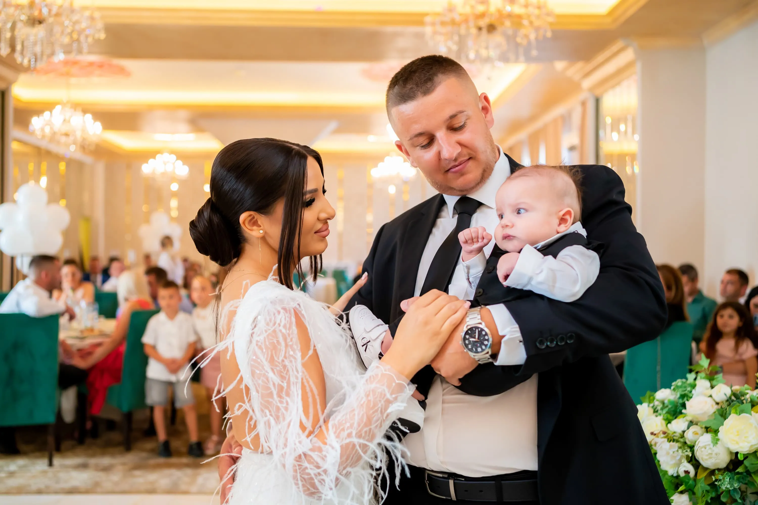Părinții țin în brațe un bebeluș îmbrăcat elegant într-un costum negru, în timpul unei ceremonii de botez. Mama poartă o rochie albă cu detalii delicate, iar tatăl poartă un costum negru. Ei sunt într-o sală de evenimente, decorată elegant, cu invitați așezați la mese în fundal. Fotografie realizată de Flaviu Studio.