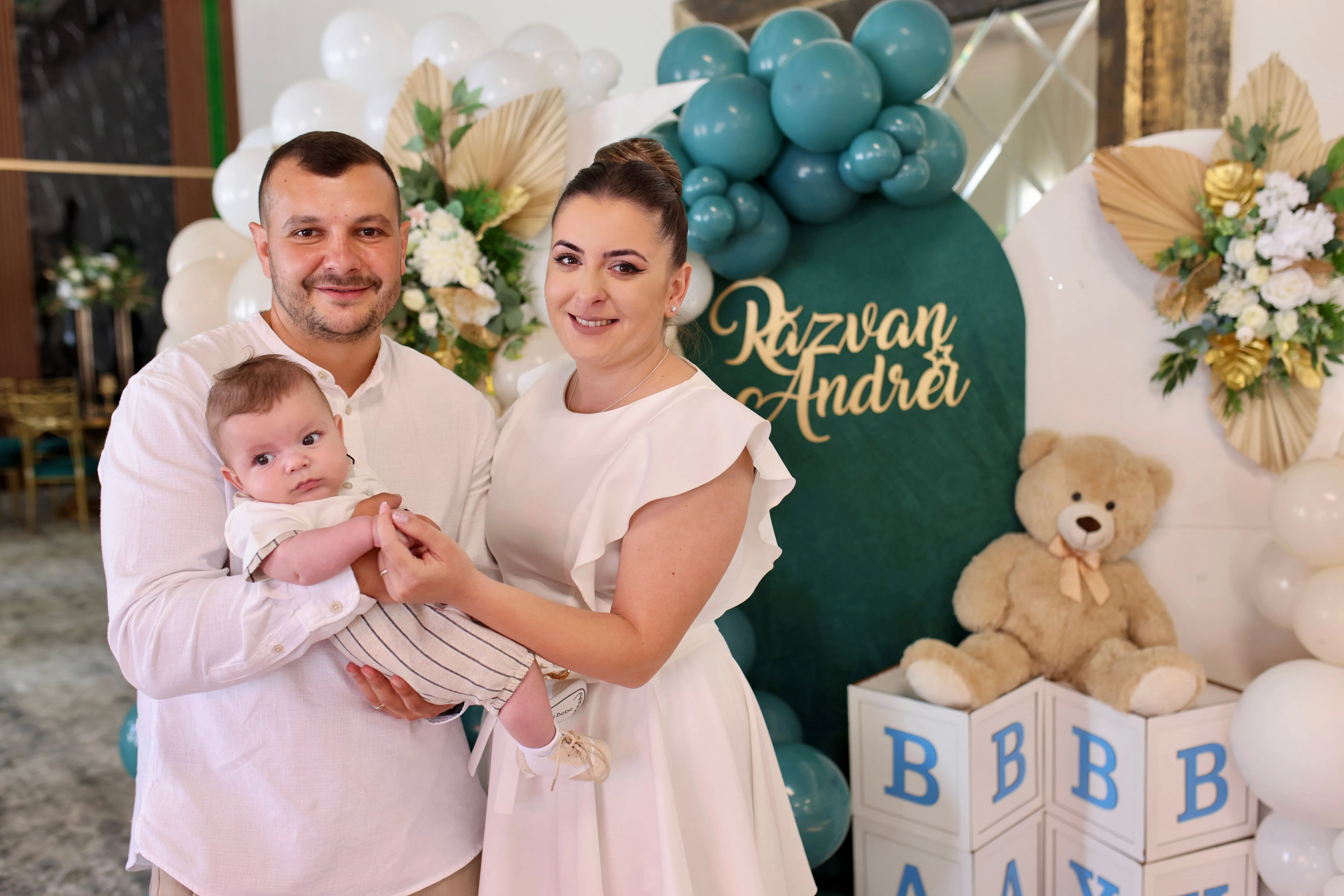 O familie fericită la botezul bebelușului lor, Răzvan Andrei. Tatăl, îmbrăcat în cămașă albă, ține în brațe bebelușul, care poartă o salopetă dungată și o cămașă albă. Mama, îmbrăcată într-o rochie albă cu volane, zâmbește spre cameră. În spatele lor se află o decorare festivă cu baloane albe și verzi, un urs de pluș și o inscripție cu numele Răzvan Andrei. Fotografia este realizată de Flaviu Studio și surprinde un moment de bucurie și familie.