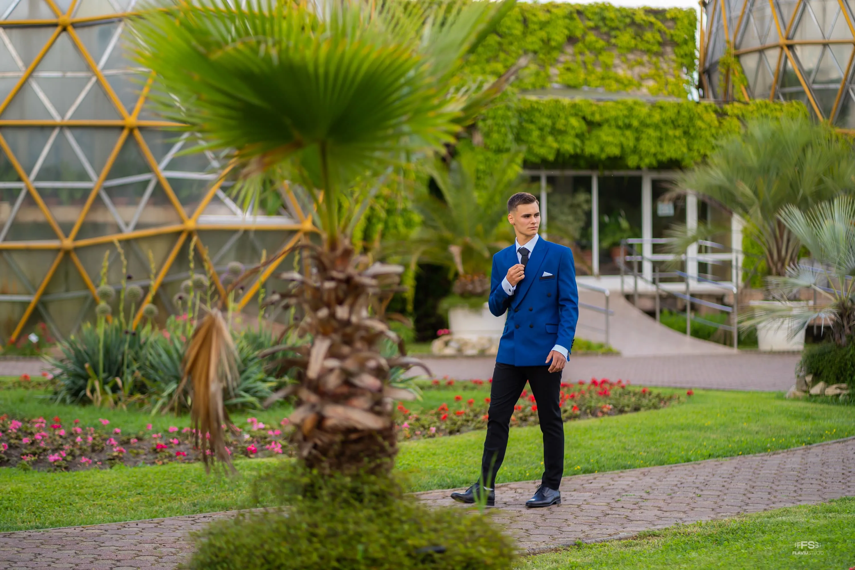 Fotografie de majorat cu Bogdan purtând un costum albastru, pozând într-o grădină luxuriantă cu arhitectură modernă în fundal. Realizată de Flaviu Studio.