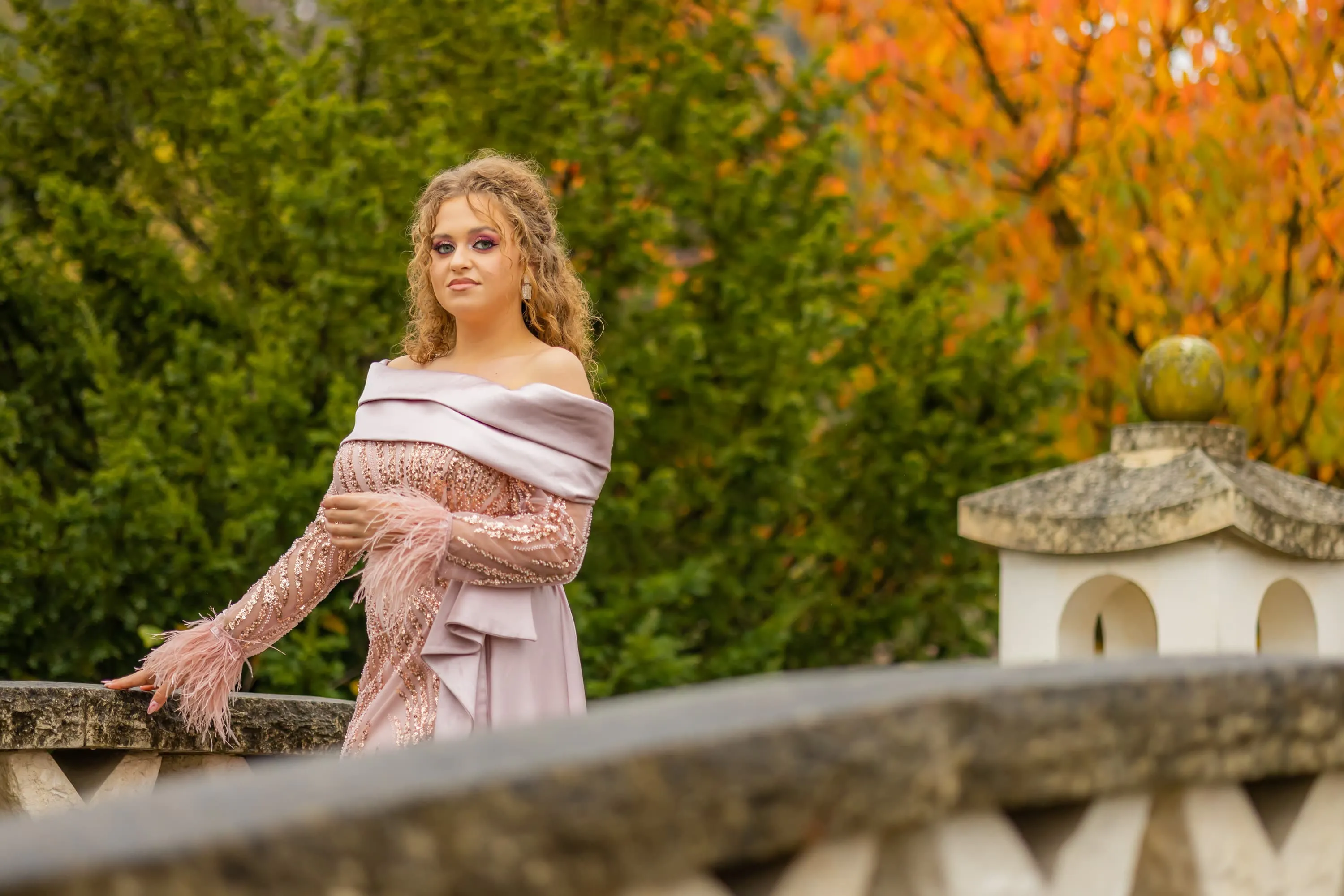 Fotografie de majorat cu Alexandra purtând o rochie roz pal cu pene, pozând pe un pod de piatră cu un fundal de copaci colorați de toamnă. Realizată de Flaviu Studio.