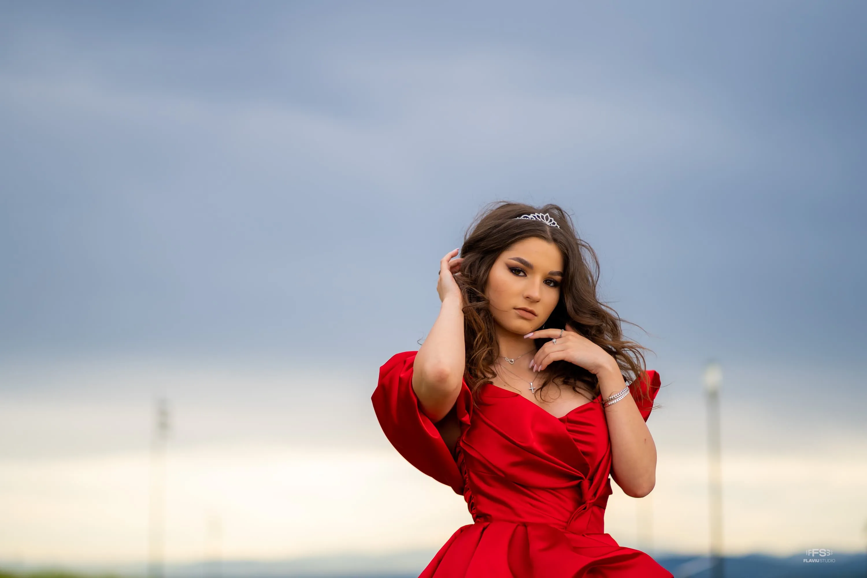 Fotografie de majorat cu Giulia purtând o rochie roșie, pozând cu mâna în păr sub un cer albastru deschis. Realizată de Flaviu Studio.