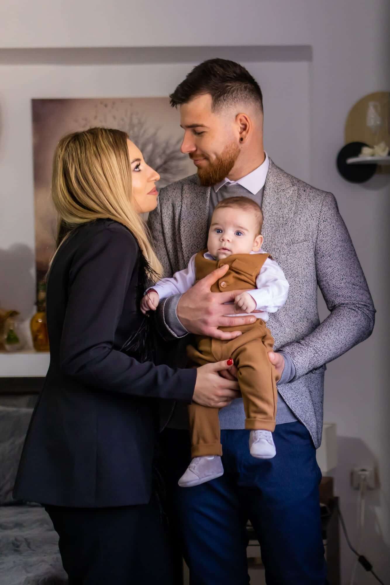 Fotografie de botez realizată de Flaviu Studio cu părinții și bebelușul Eduard-Nicolas. Tatăl, îmbrăcat într-un costum gri, ține în brațe bebelușul, iar mama, îmbrăcată într-un costum negru elegant, îi privește cu dragoste. Momentul surprinde bucuria și intimitatea familiei în timpul botezului.