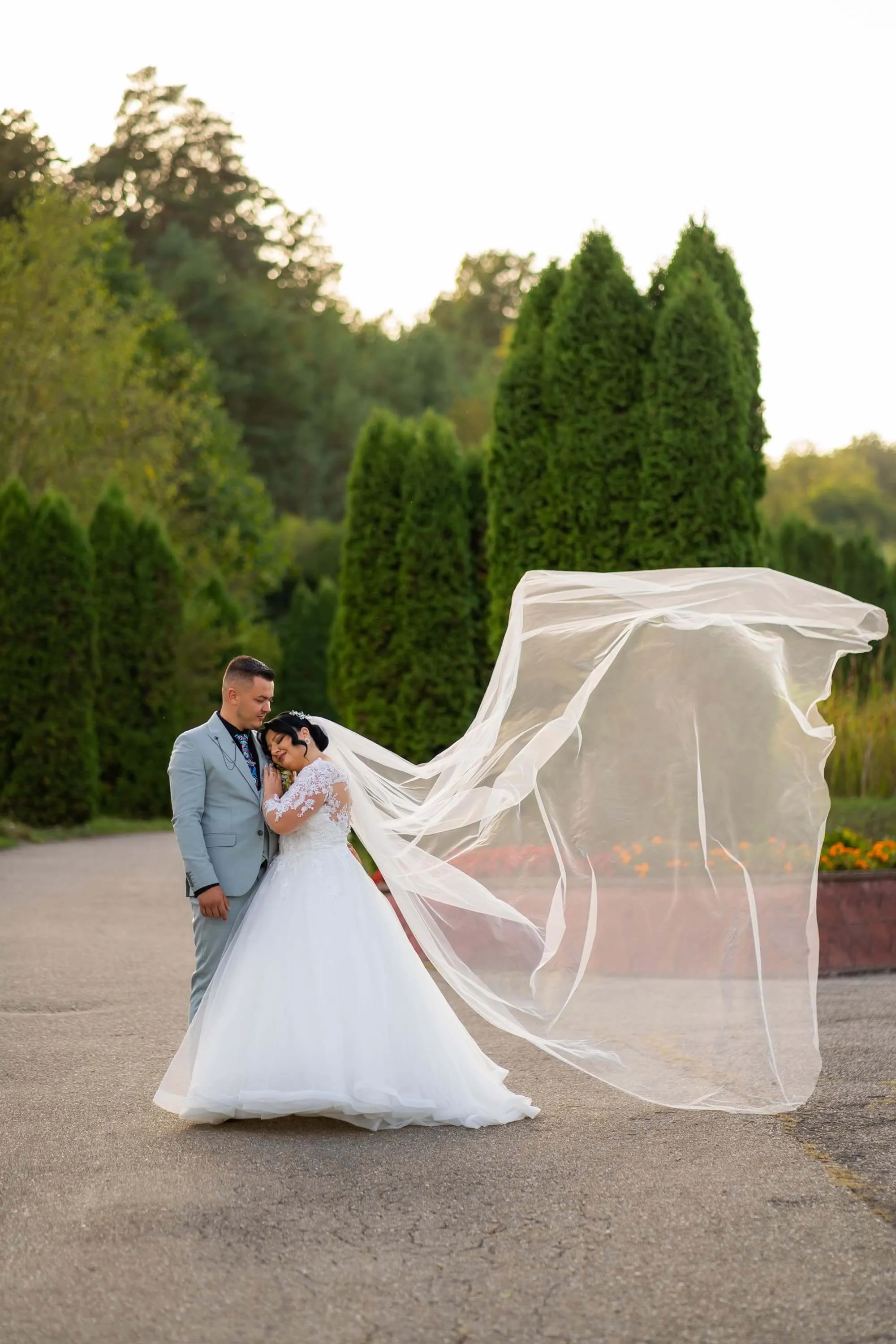 Fotografie de nuntă realizată de Flaviu Studio cu Andreea și Andrei în grădină, cu voalul miresei fluturând în vânt. Mireasa poartă o rochie albă elegantă, iar mirele este îmbrăcat într-un costum gri deschis. Momentul surprinde tandrețea și bucuria cuplului într-un cadru natural.