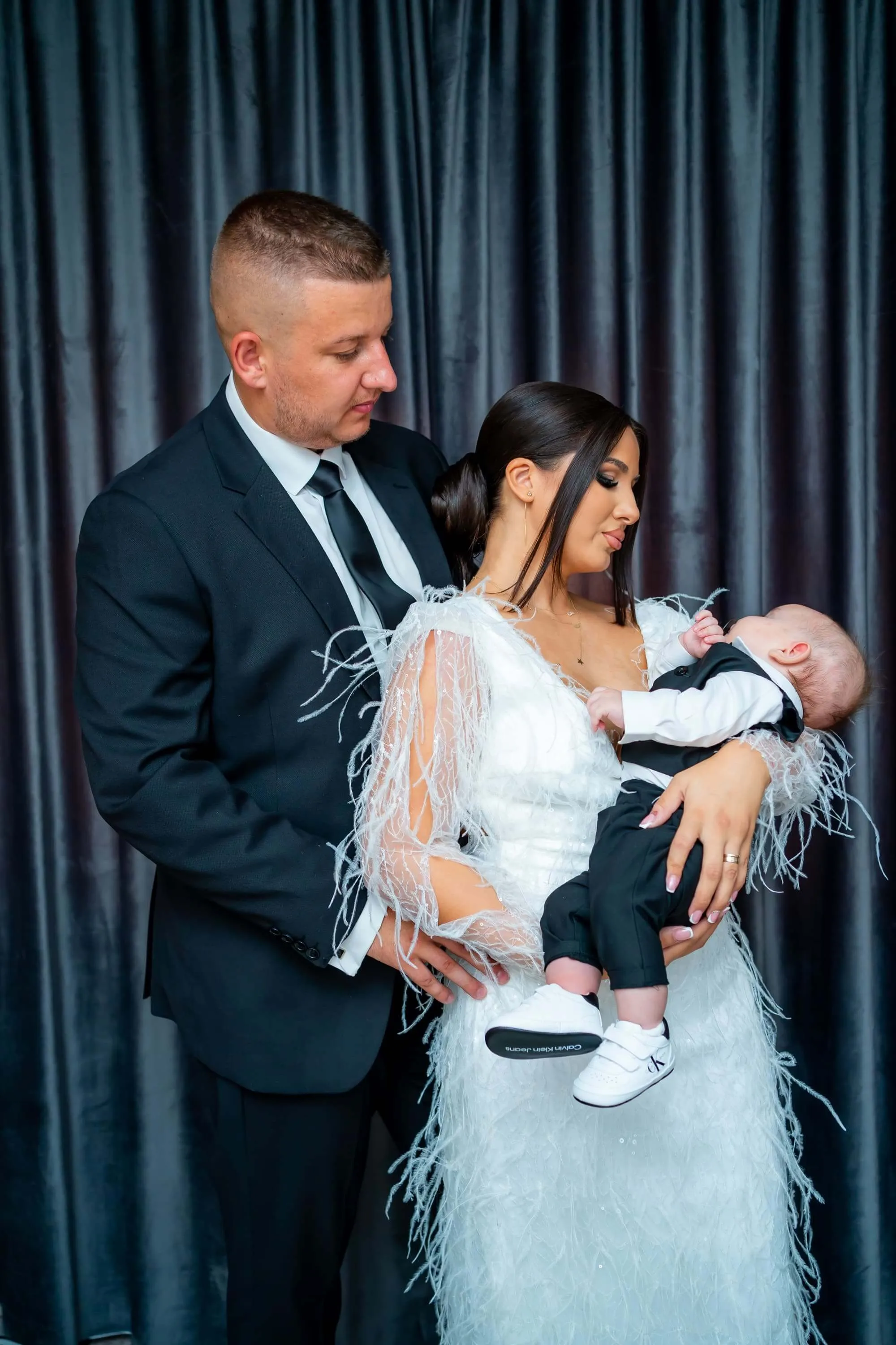 Fotografie de botez realizată de Flaviu Studio cu părinții și bebelușul Iair. Tatăl, îmbrăcat într-un costum negru, și mama, îmbrăcată într-o rochie albă cu detalii de pene, țin bebelușul îmbrăcat într-un costum alb-negru. Imaginea surprinde un moment de tandrețe și iubire familială în timpul botezului.