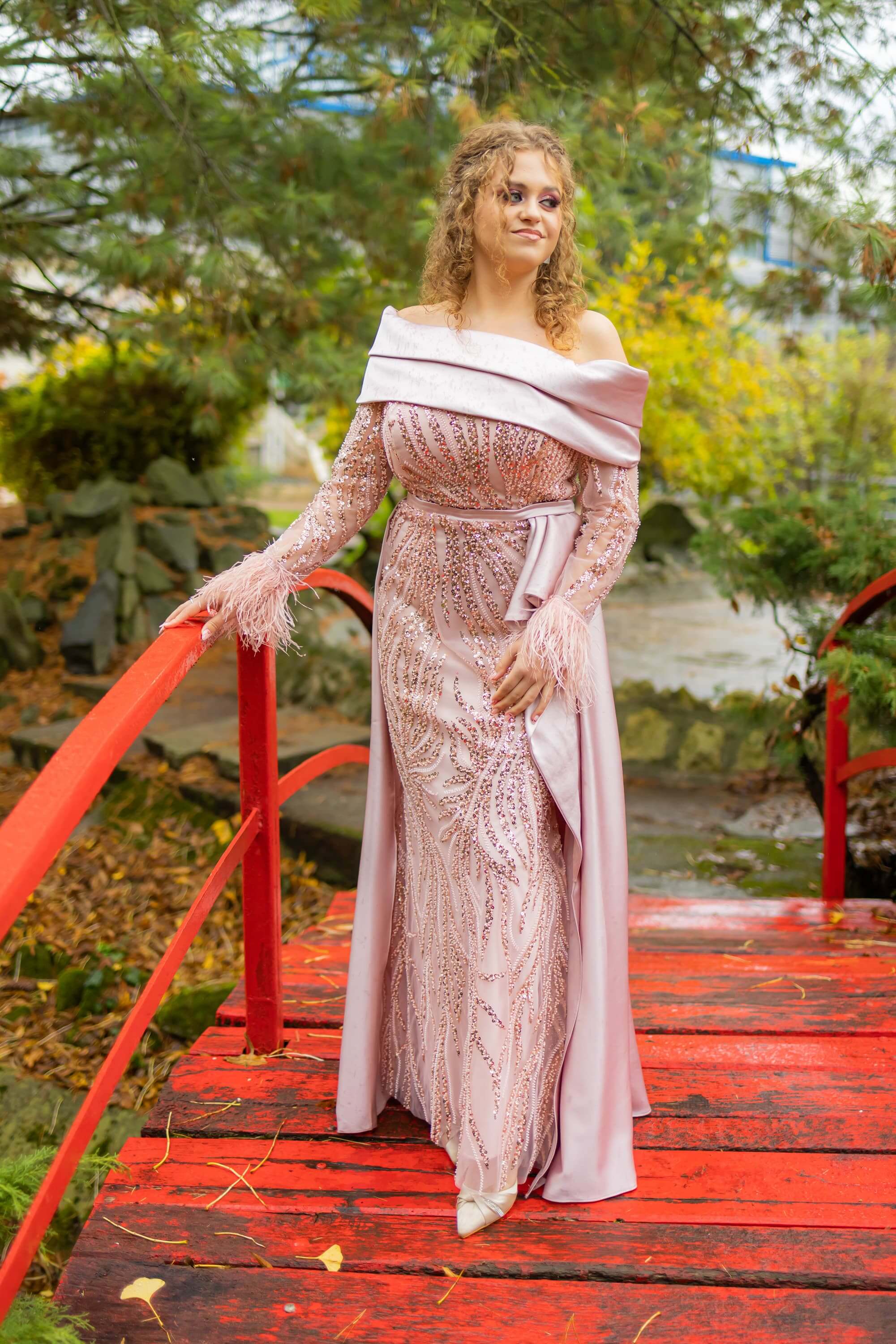 Fotografie de la majoratul Alexandrei realizată de Flaviu Studio. Alexandra poartă o rochie roz pal cu detalii strălucitoare și pene la mâneci, stând pe un pod roșu într-un cadru natural. Imaginea surprinde eleganța și frumusețea sărbătoritei într-un decor de toamnă.