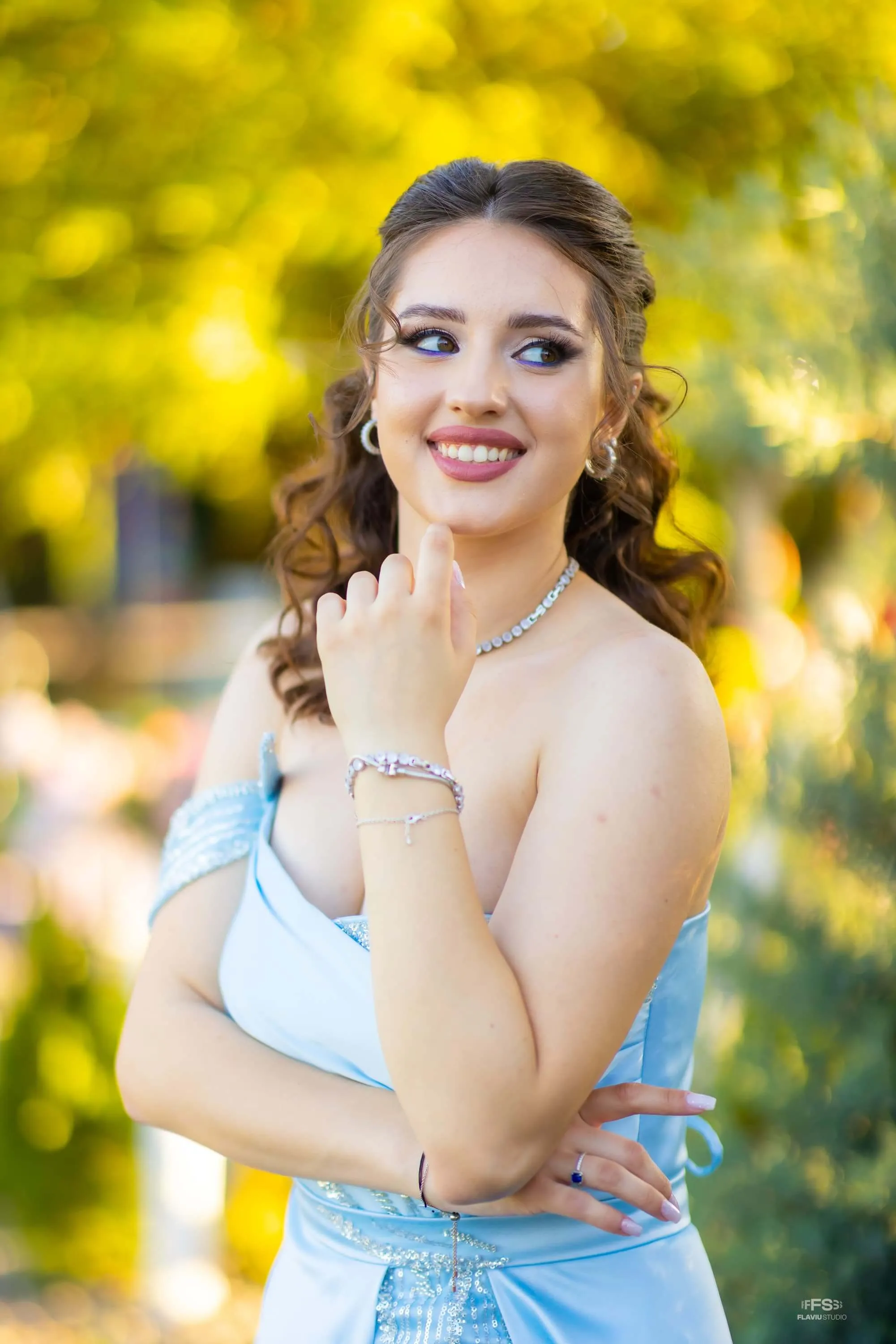 Fotografie de la majoratul Antoniei realizată de Flaviu Studio. Antonia poartă o rochie albastră elegantă și accesorii strălucitoare, zâmbind și privindu-se peste umăr. Fundalul natural, cu lumini și culori calde, completează atmosfera festivă a evenimentului.