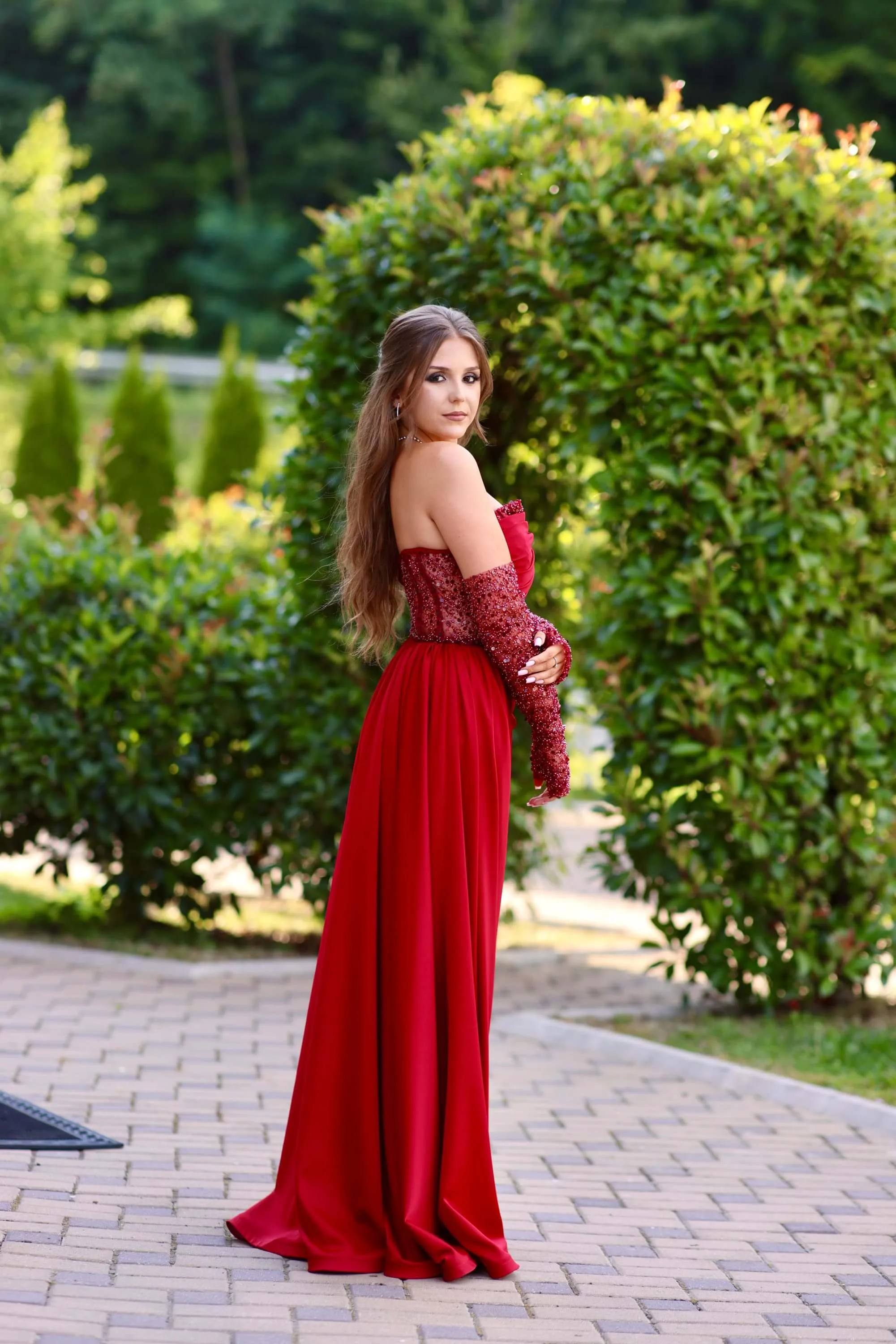 Fotografie de la majoratul Arianei realizată de Flaviu Studio. Ariana poartă o rochie roșie elegantă, cu mâneci lungi și detalii strălucitoare, stând într-un cadru natural cu verdeață. Imaginea surprinde eleganța și frumusețea sărbătoritei într-un decor de grădină.