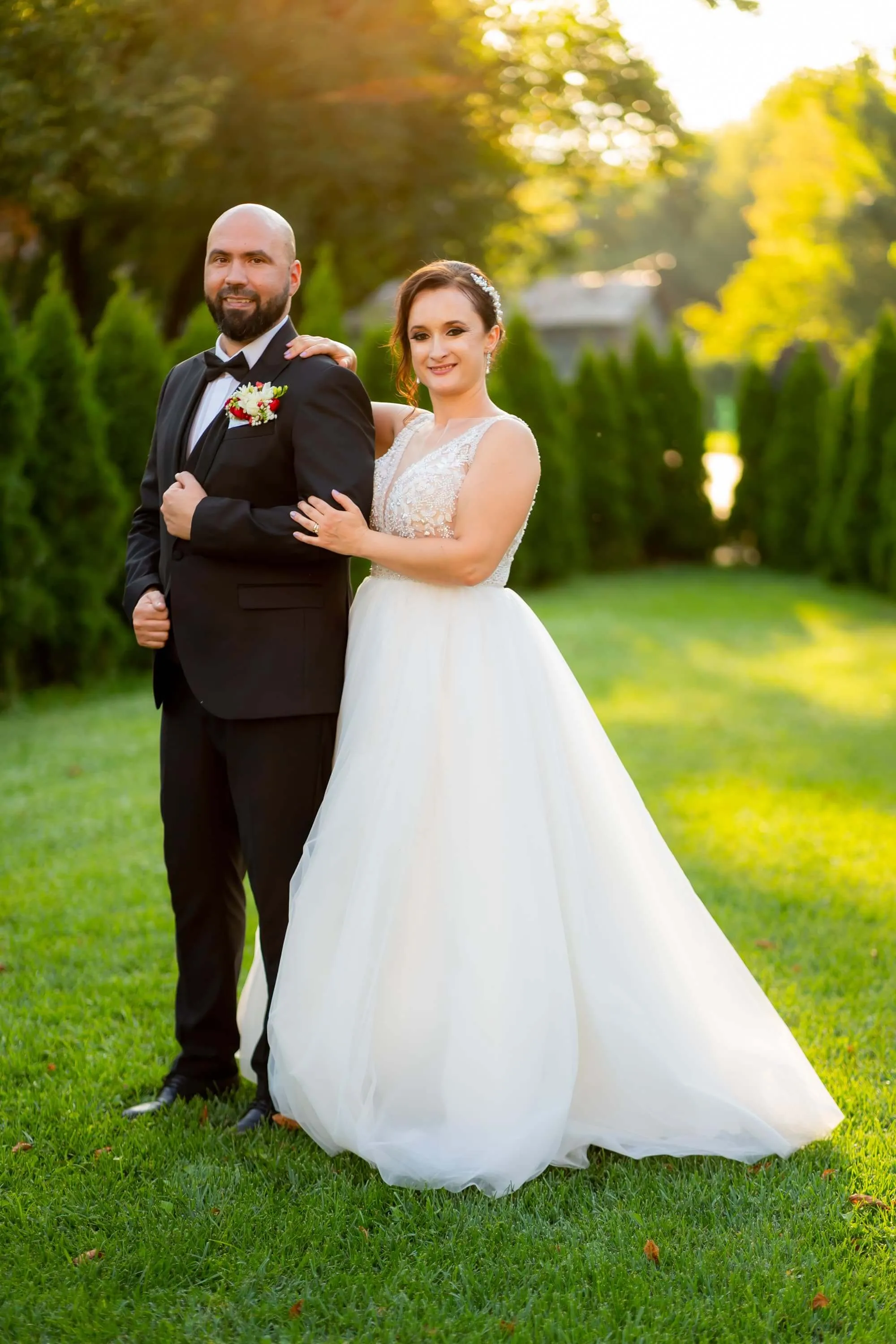 Fotografie de nuntă realizată de Flaviu Studio cu Iuliana și Mihai într-o grădină, la apus. Mireasa poartă o rochie albă elegantă cu detalii de dantelă, iar mirele este îmbrăcat într-un costum negru. Cuplul pozează elegant pe un fundal verde, luminat de soarele apusului.