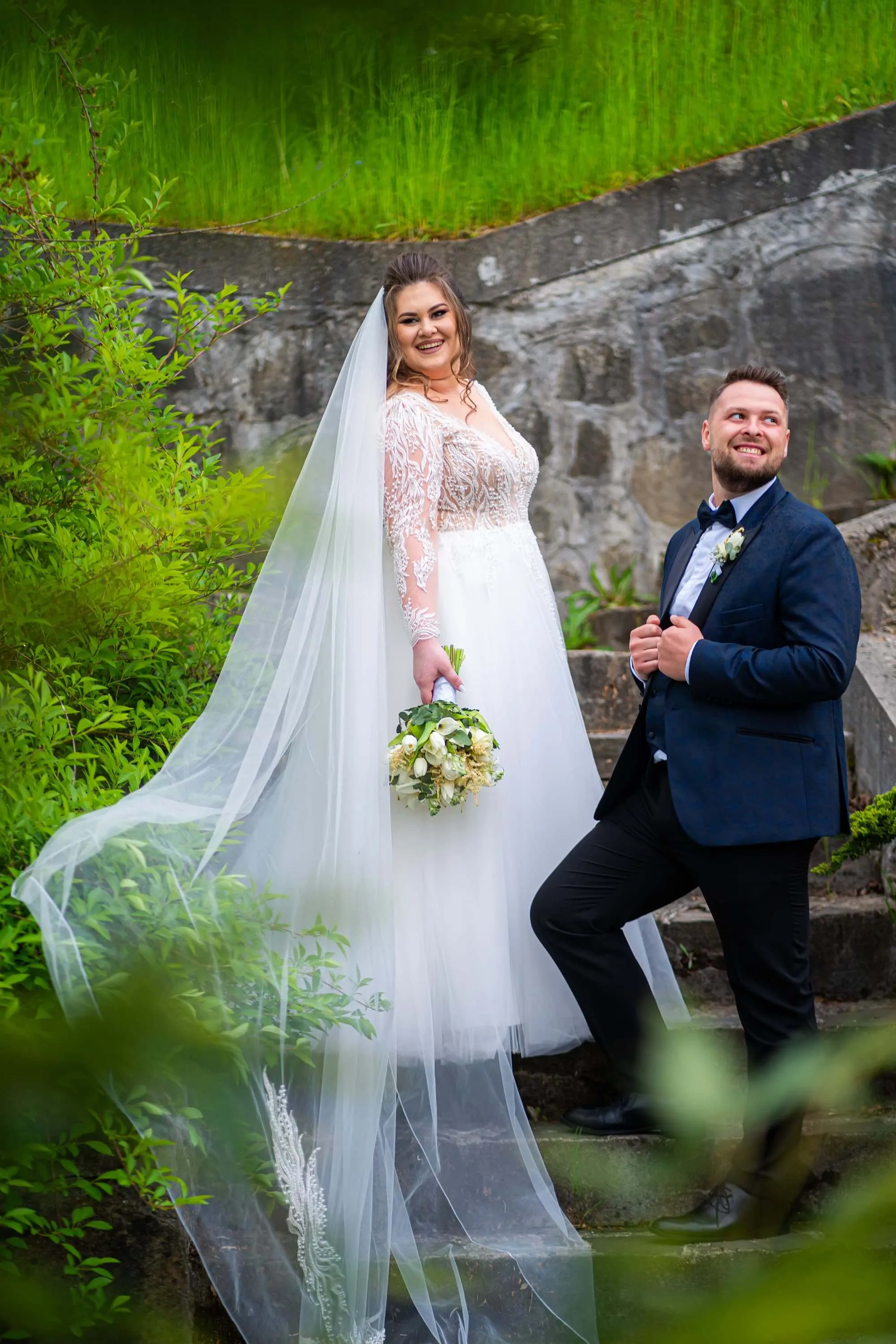 Fotografie de nuntă realizată de Flaviu Studio cu Cosmia și Robert stând pe trepte într-un cadru verde și stâncos. Mireasa poartă o rochie albă cu detalii de dantelă și un voal lung, iar mirele este îmbrăcat într-un costum elegant negru. Cuplul zâmbește fericit, surprinzându-și dragostea într-un decor natural.