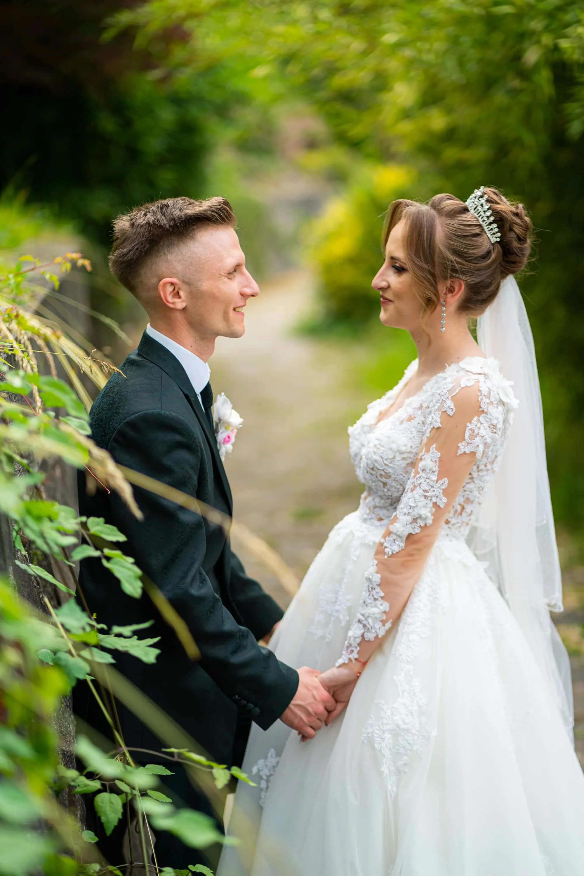 Fotografie de nuntă realizată de Flaviu Studio cu Lorena și Rareș stând față în față într-un cadru verde și romantic. Mireasa poartă o rochie albă cu detalii de dantelă și o diademă, iar mirele este îmbrăcat într-un costum negru. Momentul surprinde dragostea și bucuria cuplului într-un decor natural.